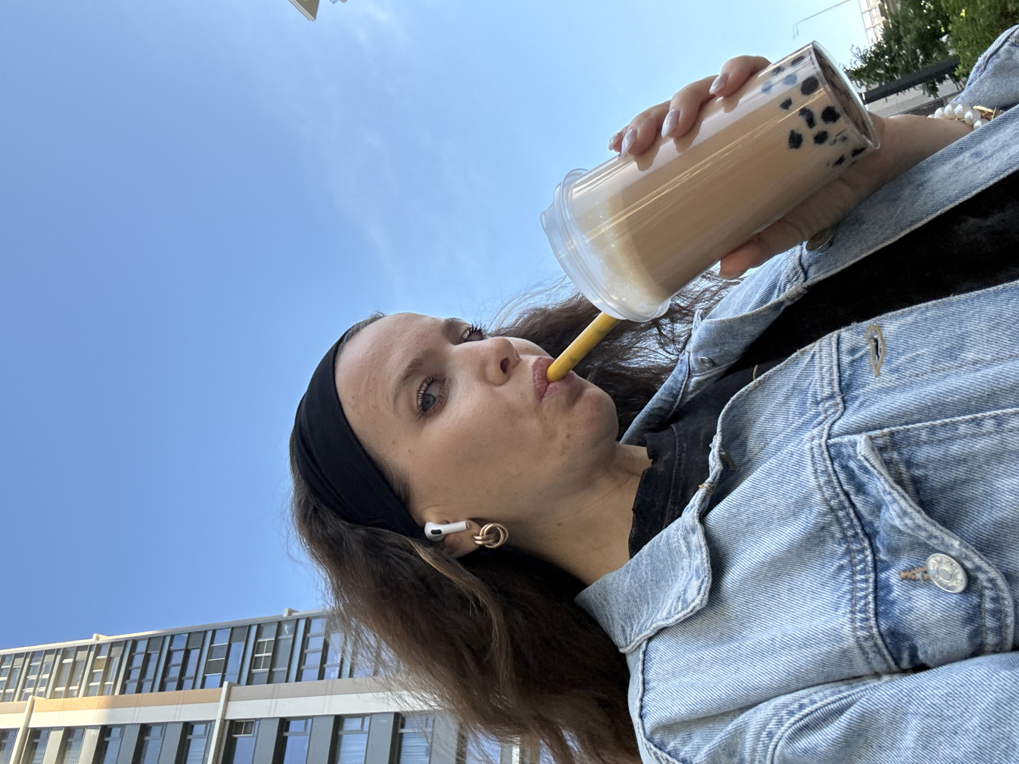 A low-angle shot shows a woman drinking a bubble tea outdoors, with a clear blue sky and a modern building in the background. The woman has long brown hair and is wearing a black headband and a denim jacket. She has a pair of white wireless earbuds in her ears and gold hoop earrings. The bubble tea is in a clear plastic cup with a yellow straw and black tapioca pearls. The woman is looking towards the camera with a slight smile. The scene appears to be set in daylight, possibly in the afternoon, given the bright sky and sunlight. The architecture of the building suggests an urban setting, which is consistent with the Moscow, Russia location context. The overall mood is casual and relaxed.