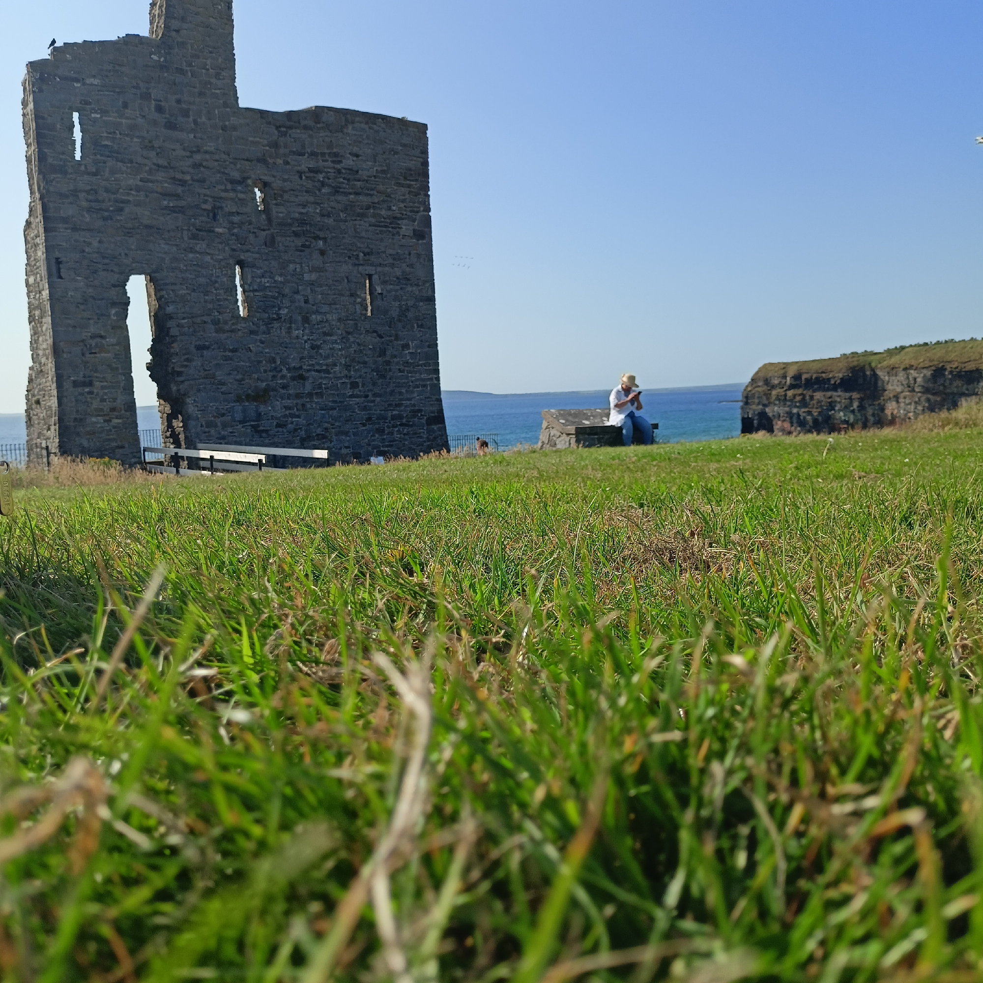 In Ballybunion, Ireland, a stone ruin stands against a clear blue sky. The ruin is made of dark grey stones and has large openings. In the foreground, a grassy field slopes upwards, with blades of grass appearing in sharp focus. In the mid-ground, a person wearing a straw hat, a white shirt, and blue jeans sits on a stone bench, looking at their phone. The sea stretches out behind them, and on the right, a rocky cliff face rises from the water.