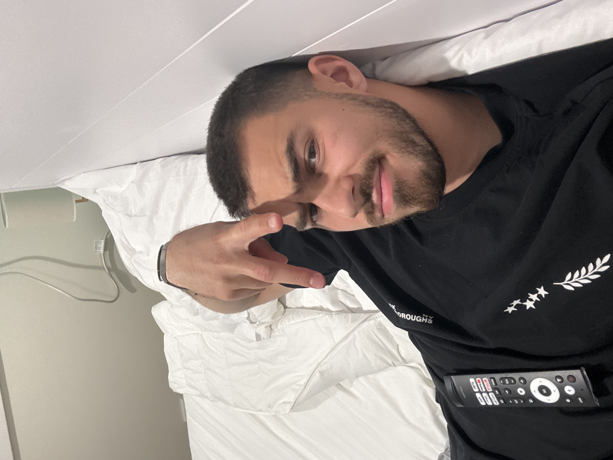 A man with short dark hair and a beard is lying in a bed covered in white sheets, possibly in a hotel room in Hampton, United States of America. He is wearing a black t-shirt and holding a TV remote. He is making a hand gesture, possibly a peace sign. The background includes a wall and a power outlet.