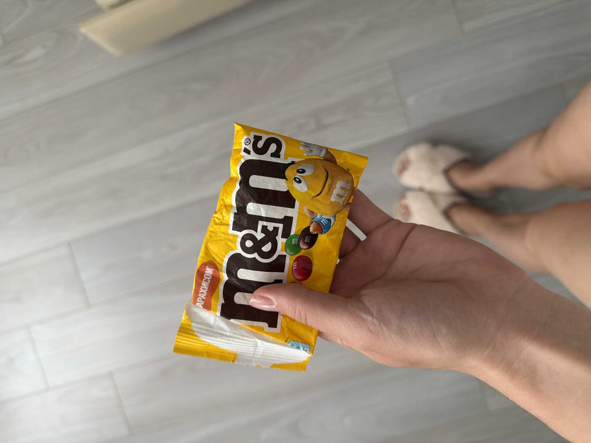 A person's hand is holding a yellow M&M's candy package. The package has the M&M's logo and images of M&M's candies. The person is wearing fluffy slippers and their legs are visible in the background. The scene appears to be indoors, on a light-colored wooden floor. This image was taken in Semiluki, Russia.