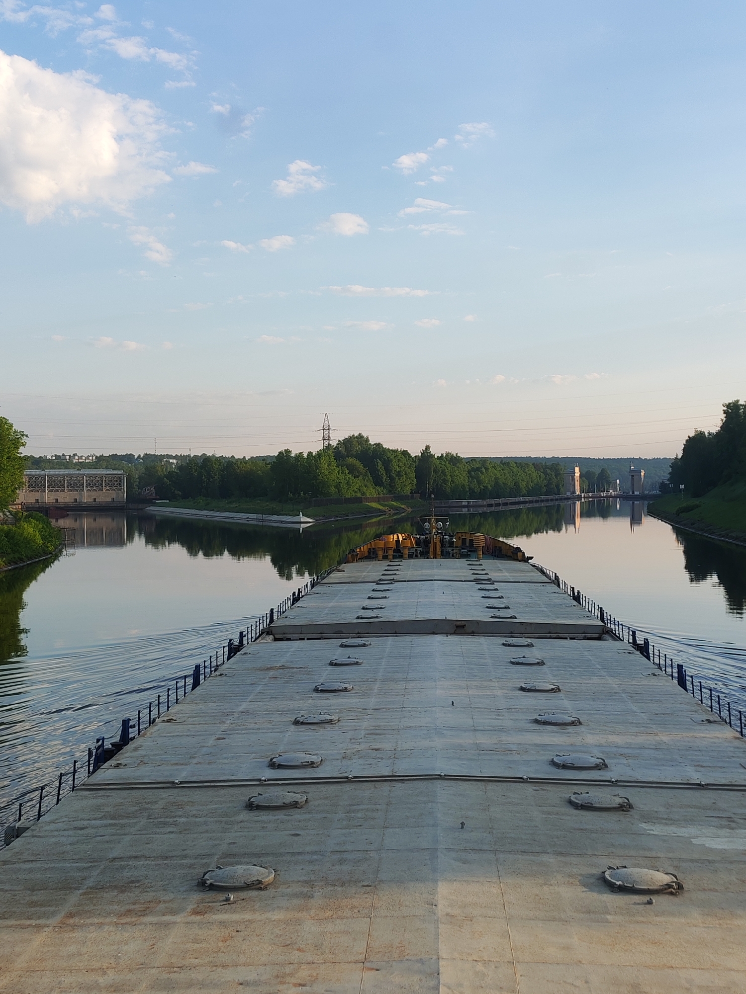 View from the deck of a barge traveling on a canal in посёлок опытного хозяйства «Ермолино», Russia. The barge deck is flat and grey with dark round hatches. The water is calm and reflects the sky and the green trees lining the canal banks. In the distance are structures, including what appear to be towers and a building. Power lines stretch across the scene above the trees. The sky is blue with scattered white clouds.