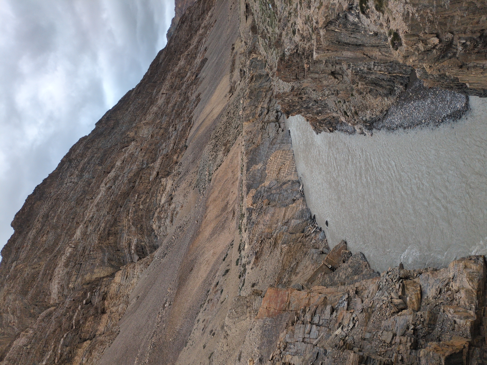 The image depicts a stark, mountainous landscape with a river flowing through a gorge. The terrain is characterized by rugged, stratified rock formations in shades of brown and tan, with scree slopes leading up to a heavily clouded sky. The river is wide and appears to be carrying sediment, giving it a murky, grayish-brown color. The banks of the gorge are steep and rocky, with some sparse vegetation clinging to the upper slopes. The overall impression is one of a remote and somewhat desolate environment. The image does not contain any individuals, actions, or interactions. The location provided, Dhar Sajam, India, is a city; however, the scene appears to be a natural geographical feature, possibly a river valley within a mountainous region of India, rather than an urban setting.