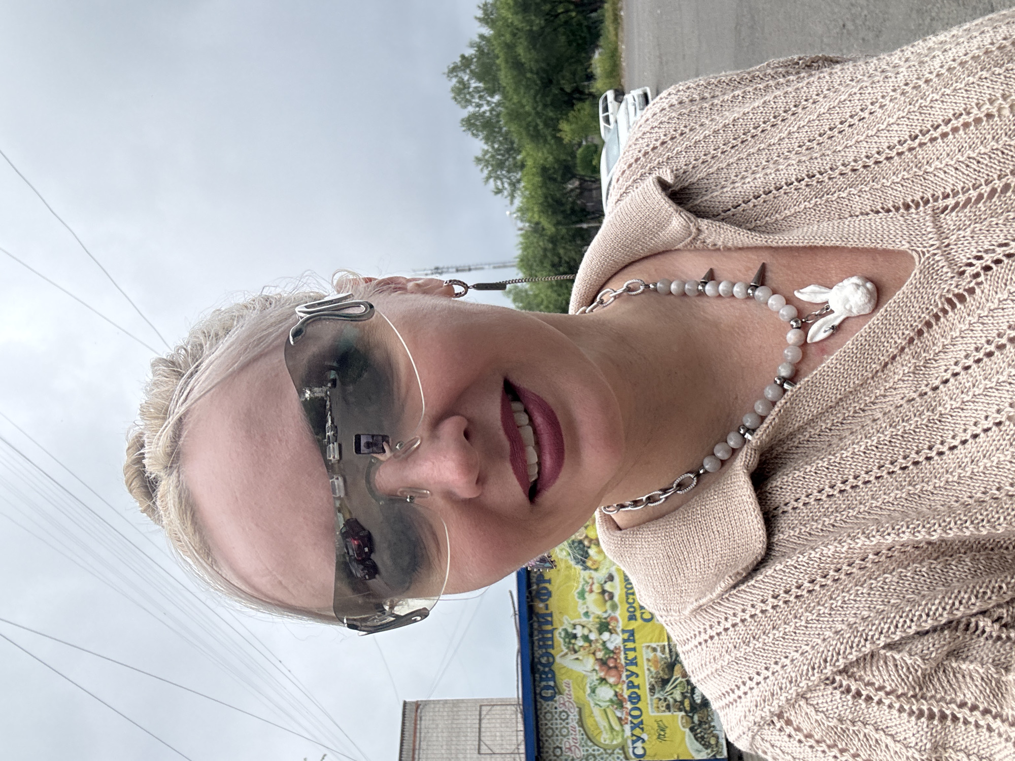 A blonde woman with her hair pulled back in a bun is taking a selfie outdoors. She is wearing large, reflective sunglasses, a light-colored knitted shirt, and a necklace with a bunny charm. In the reflection of her sunglasses, a smartphone is visible, indicating she is holding a phone to take the picture. Behind her, a yellow and blue banner with Cyrillic text that appears to be advertising "ОВOЩИ-ФРУКТЫ" (Vegetables-Fruits) is displayed on the side of a vehicle. In the background, trees and a cloudy sky are visible. The scene appears to be in a parking lot in Komsomolsk-on-Amur, Russia, suggested by the Cyrillic text on the banner and the overall urban setting.