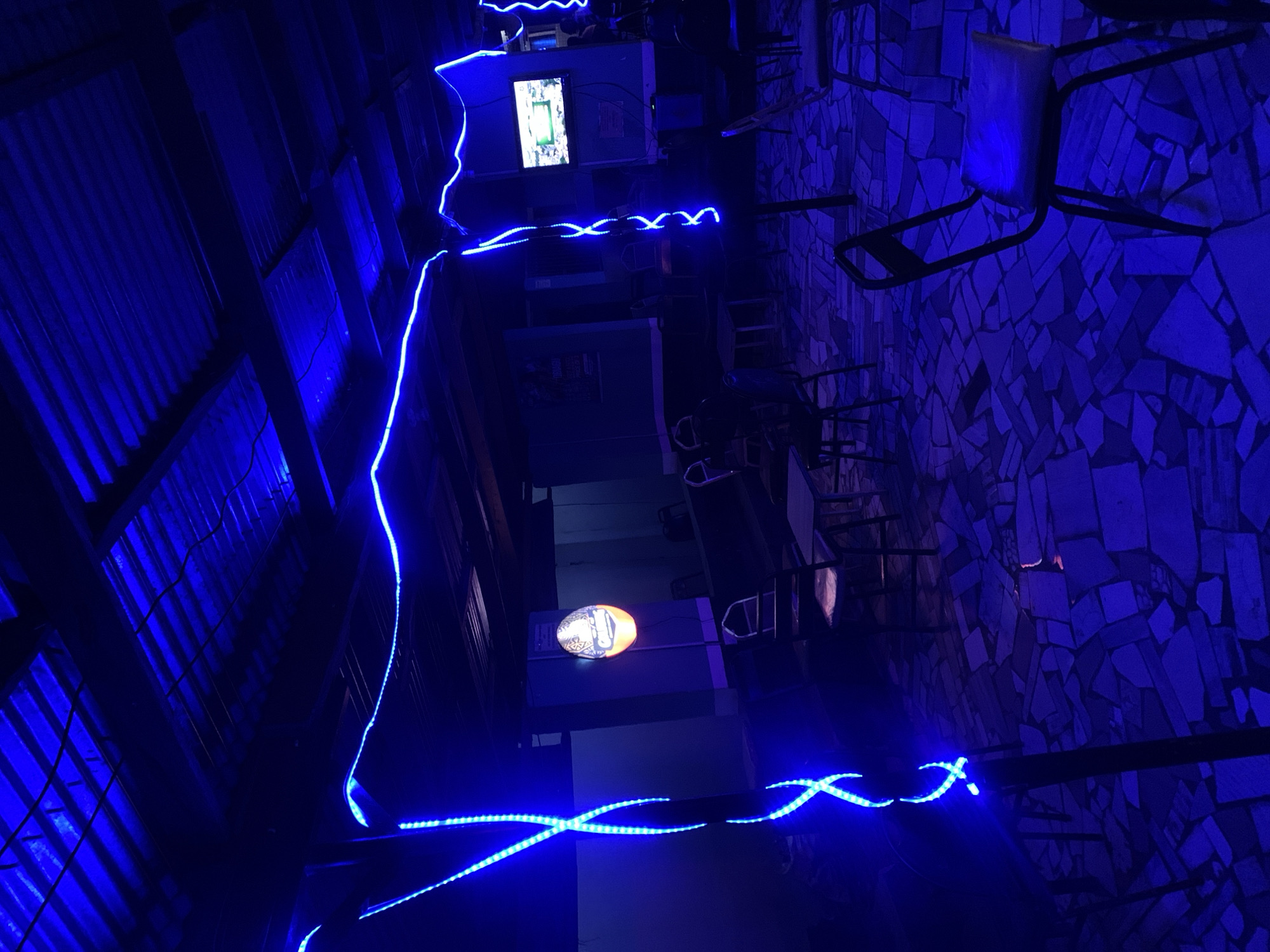 The image displays the interior of an establishment, likely a bar or lounge, characterized by intense blue lighting that gives it a distinct nighttime ambiance. The scene is devoid of people, suggesting a quiet moment or off-hours.
To the left, a corrugated metal ceiling or wall panel is heavily illuminated by bright blue LED strips. These strips snake across the entire frame, creating luminous lines and patterns, including distinct 'X' shapes in the foreground.
In the mid-ground, a monitor screen is visible, displaying an indistinct, bright image or video. Below this, a warm, golden-hued circular sign or light fixture stands out against the blue backdrop. It features an unidentifiable logo or text, possibly for a beverage brand.
The right side of the image showcases a wall with a unique, textured pattern, possibly made of mosaic or irregularly shaped tiles, also bathed in the blue light. Scattered throughout the space are dark tables and chairs, some neatly arranged or stacked, typical of a dining or social venue. The contrasting blue and golden lights, combined with the structural elements like corrugated metal and textured walls, create a modern and atmospheric setting. The entire scene points to an indoor location, likely during the evening or night. No clearly legible text is visible. The location context is New Stadium, Nigeria.