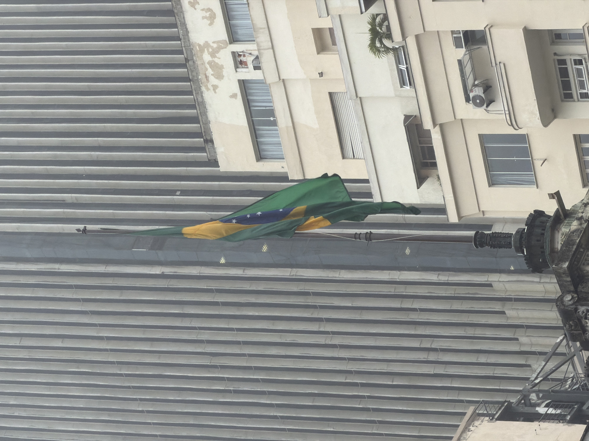 The image shows a Brazilian flag flying in front of a large, modern building with a ribbed facade in Rio de Janeiro, Brazil. The flag, predominantly green and yellow with a blue sphere and white stars, is suspended from a flagpole. Below the modern building, a lighter-colored, older building with multiple windows and balconies is visible. The sky appears overcast. The scene depicts an urban environment with a mix of architectural styles.