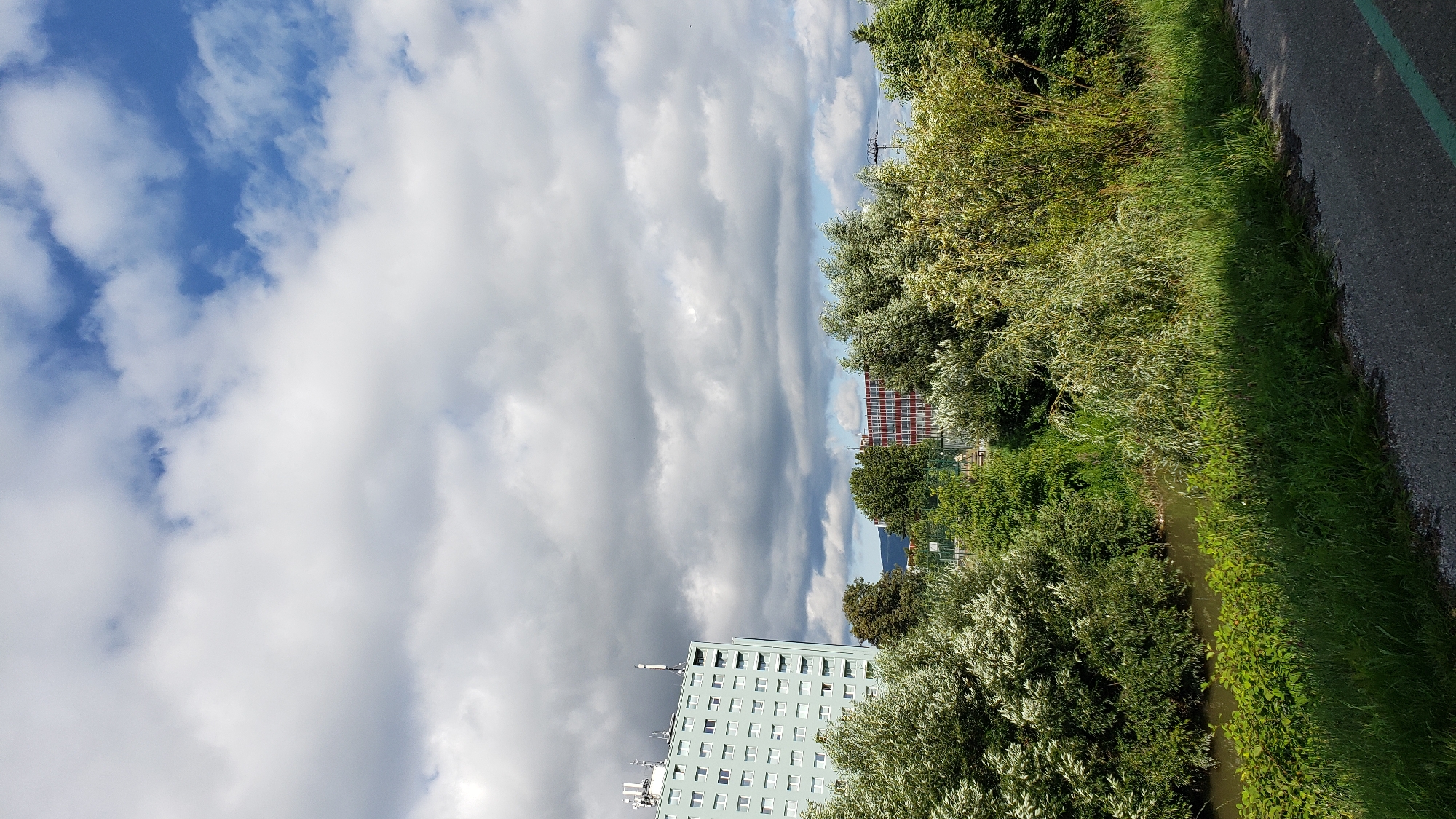 The image captures a view of a city on a cloudy day. On the left, a tall, light blue, multi-story building with numerous windows is visible, characterized by its rectangular shape and antennas on the roof. To the right of this building, in the middle ground, is a smaller, reddish-brown building with a grid-like pattern of windows, possibly a commercial or industrial structure. The foreground is dominated by lush green foliage, including trees and bushes, and a narrow body of water that appears to be a river or stream. A paved pathway, possibly a road or sidewalk, runs along the bottom right of the image, marked with a faint green line. The sky is filled with a dramatic display of clouds, ranging from bright white to dark gray, interspersed with patches of blue sky. The scene suggests an urban or semi-urban environment, likely on the outskirts of a city, given the presence of both buildings and significant natural vegetation. Based on the architecture of the tall building, it resembles a residential apartment block. The overall impression is that of a somewhat overcast day in an area where nature meets urban development. This scene is consistent with areas within the city of Prešov, Slovakia, where such juxtapositions of modern buildings and natural waterways are common.