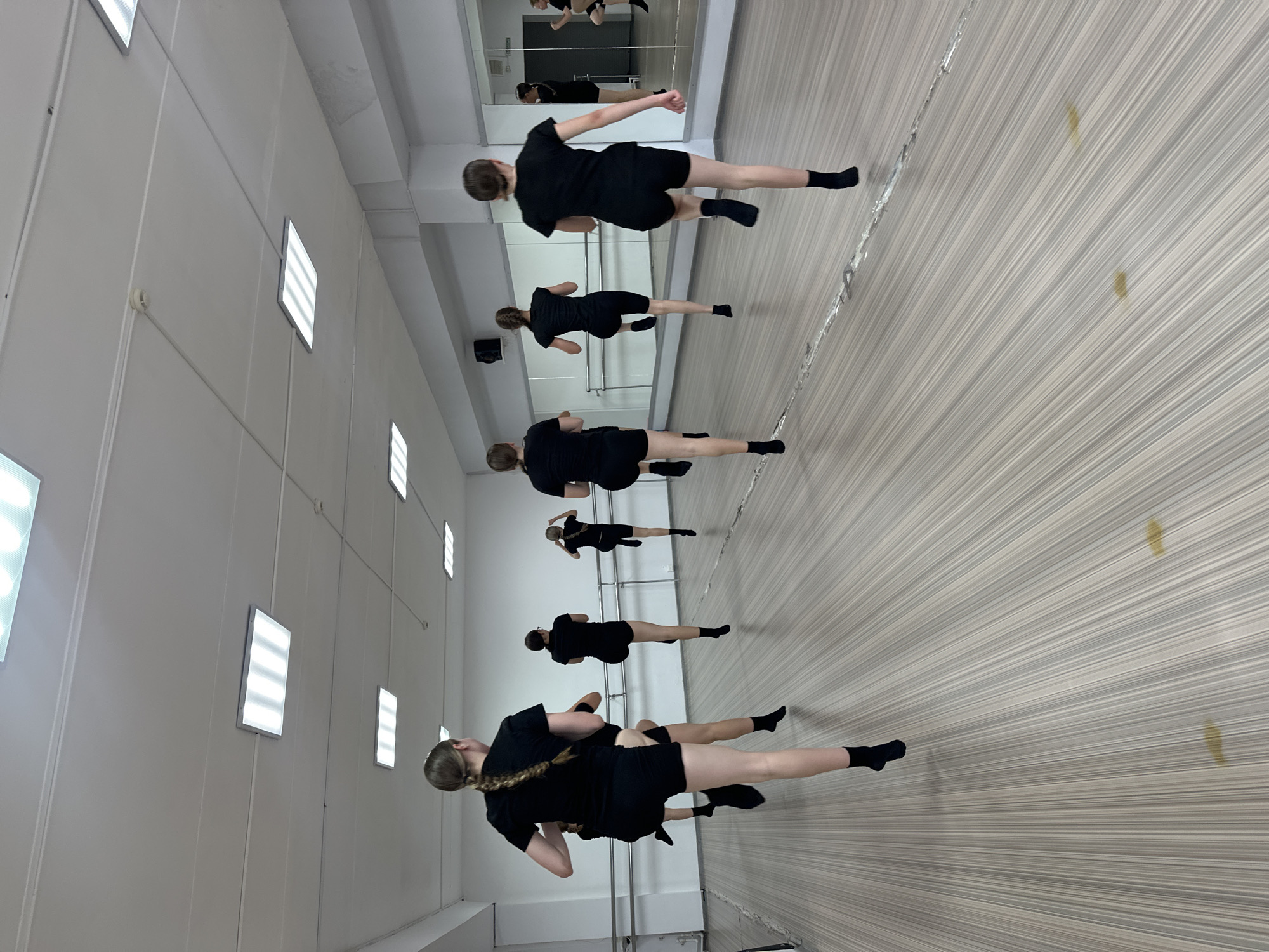 In a dance studio, possibly located in Votkinsk, Russia, a group of seven young women are engaged in a synchronized high-knee exercise. They are all dressed in black, form-fitting attire consisting of shorts and t-shirts, and are wearing black socks. Their faces are not visible as the image is taken from behind. The room features a light-colored wooden floor with a striped pattern, white walls, and a mirrored wall on the right side of the frame, reflecting some of the participants and the studio's interior. Fluorescent lighting panels are integrated into the white ceiling. A barre runs along the wall opposite the mirror, suggesting a dedicated space for dance or gymnastics. Some minor, yellowish stains are visible on the floor in the foreground. The participants are captured mid-action, with one leg raised high in front of them.