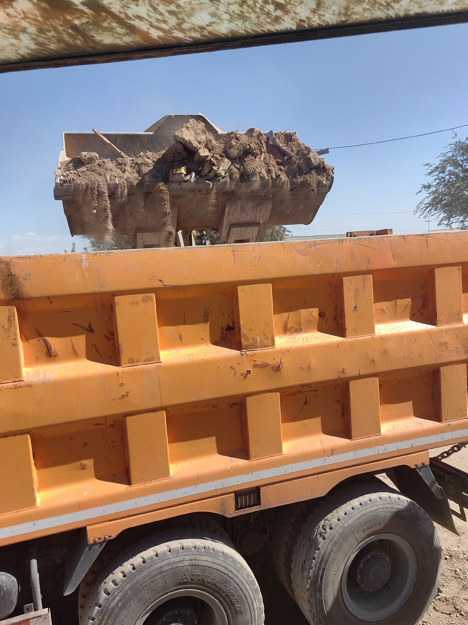 The image shows a side view of a large orange dump truck, likely in Shymkent, Kazakhstan. The truck's bed is filled with dirt and rocks, and it appears to be in the process of being loaded or unloaded. Above the truck, a shovel or scoop is visible, also filled with dirt. The sky is clear and blue, suggesting a sunny day. The ground is covered with dirt and gravel, consistent with a construction site or a similar environment. The truck's tires are large and robust, indicating its heavy-duty purpose. The overall scene depicts an industrial or construction activity in an outdoor setting.