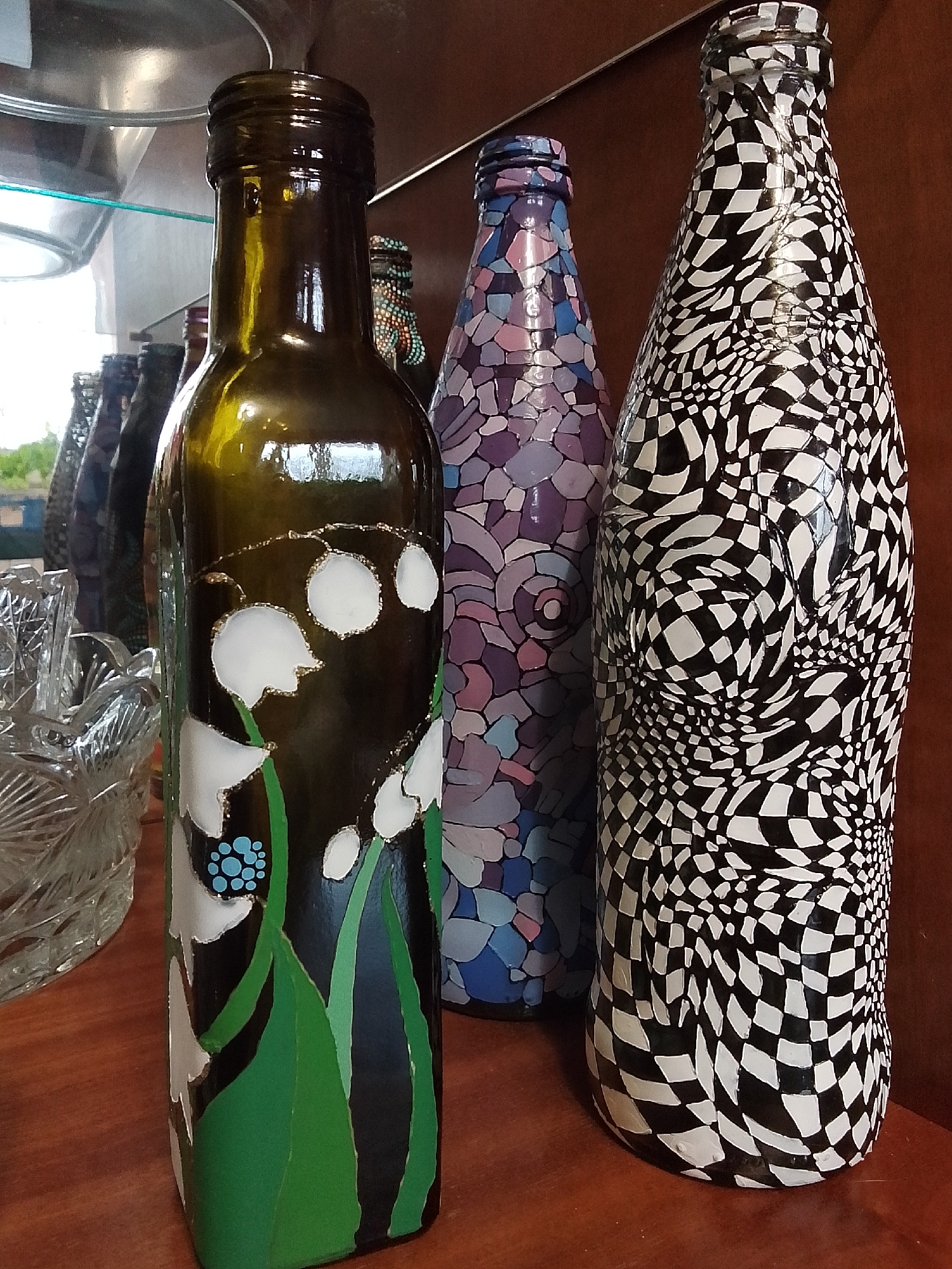 The image captures a collection of hand-decorated bottles arranged on a wooden surface, likely a shelf, in an indoor setting. In the foreground, a dark green bottle is adorned with white lily-of-the-valley flowers, outlined in gold, complemented by green leaves and a cluster of small blue dots. To its right, a bottle features a mosaic-like pattern in shades of purple, blue, and pink. The most prominent bottle on the far right displays a striking black and white checkerboard pattern that appears to swirl and distort, creating an optical illusion.
Several other decorative bottles are partially visible in the background. To the left of the green bottle, a clear, ornate cut-glass bowl or dish adds to the display. The background is a dark brown, possibly wood paneling, with a reflective surface above, suggesting a glass shelf or ceiling. Natural light from the left casts reflections and highlights on the glossy surfaces of the bottles and glassware. No activity is taking place, presenting a still life arrangement of craft items. There is no visible text. The scene appears to be set during the daytime due to the natural light.