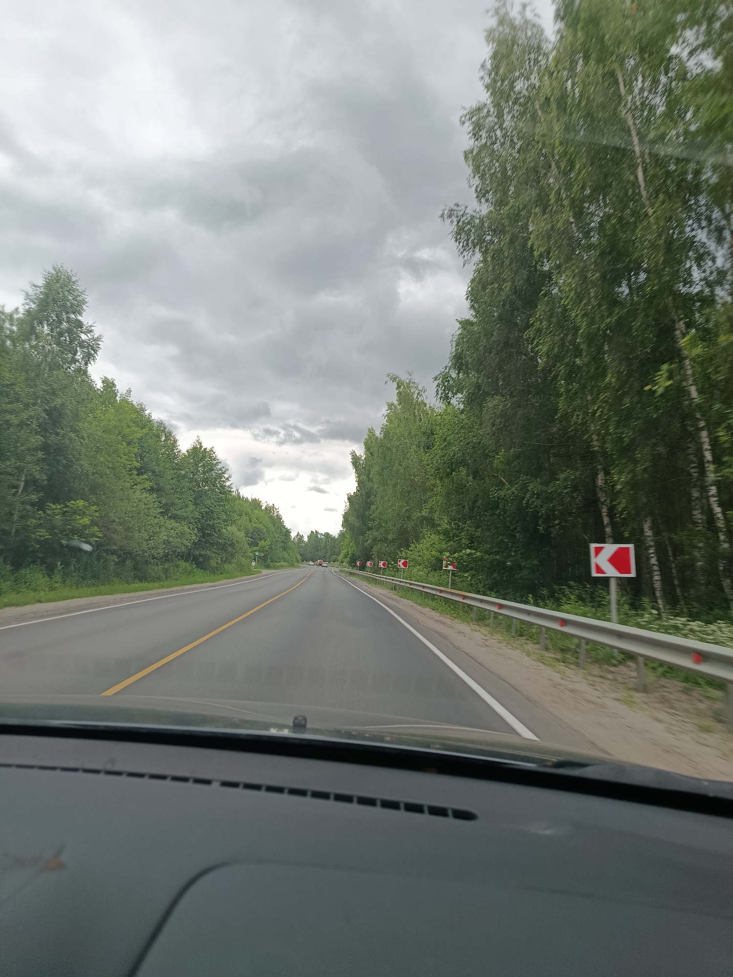 This image depicts a view from inside a car driving on a two-lane road lined with dense green trees on both sides. The sky is overcast with thick gray clouds, suggesting a cloudy or possibly rainy day. The road has a yellow line down the center and a white line marking the edge. Guardrails are visible along the right side of the road, with red markers and directional signs indicating a curve to the left. Further down the road, other vehicles can be seen in the distance, indicating normal traffic flow. The dashboard of the car is visible in the foreground, along with part of the windshield. The scene conveys a sense of travel through a rural or forested area, likely during the daytime. There is no visible text in the image.