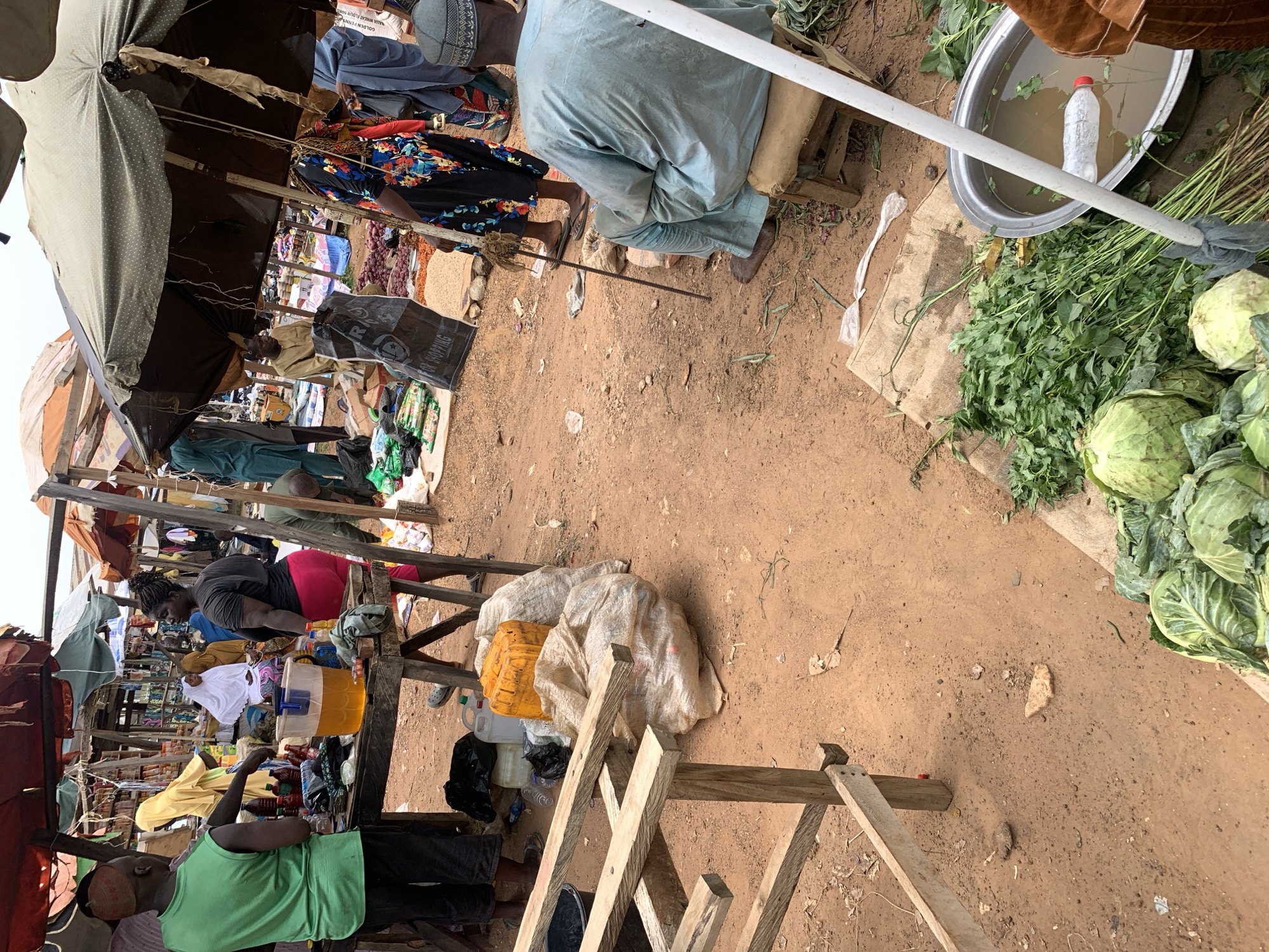 The image depicts a bustling marketplace scene in Keffi, Nigeria. Several vendors are present, sitting or standing behind makeshift wooden stalls covered with various goods. Clothing, food items, and other merchandise are visible. People are seen interacting and making transactions. The ground is predominantly dirt, with scattered debris and objects. In the foreground, there are cabbages and green leafy vegetables alongside a tub of water containing a plastic bottle. The scene is shaded by umbrellas and makeshift coverings erected over the stalls.