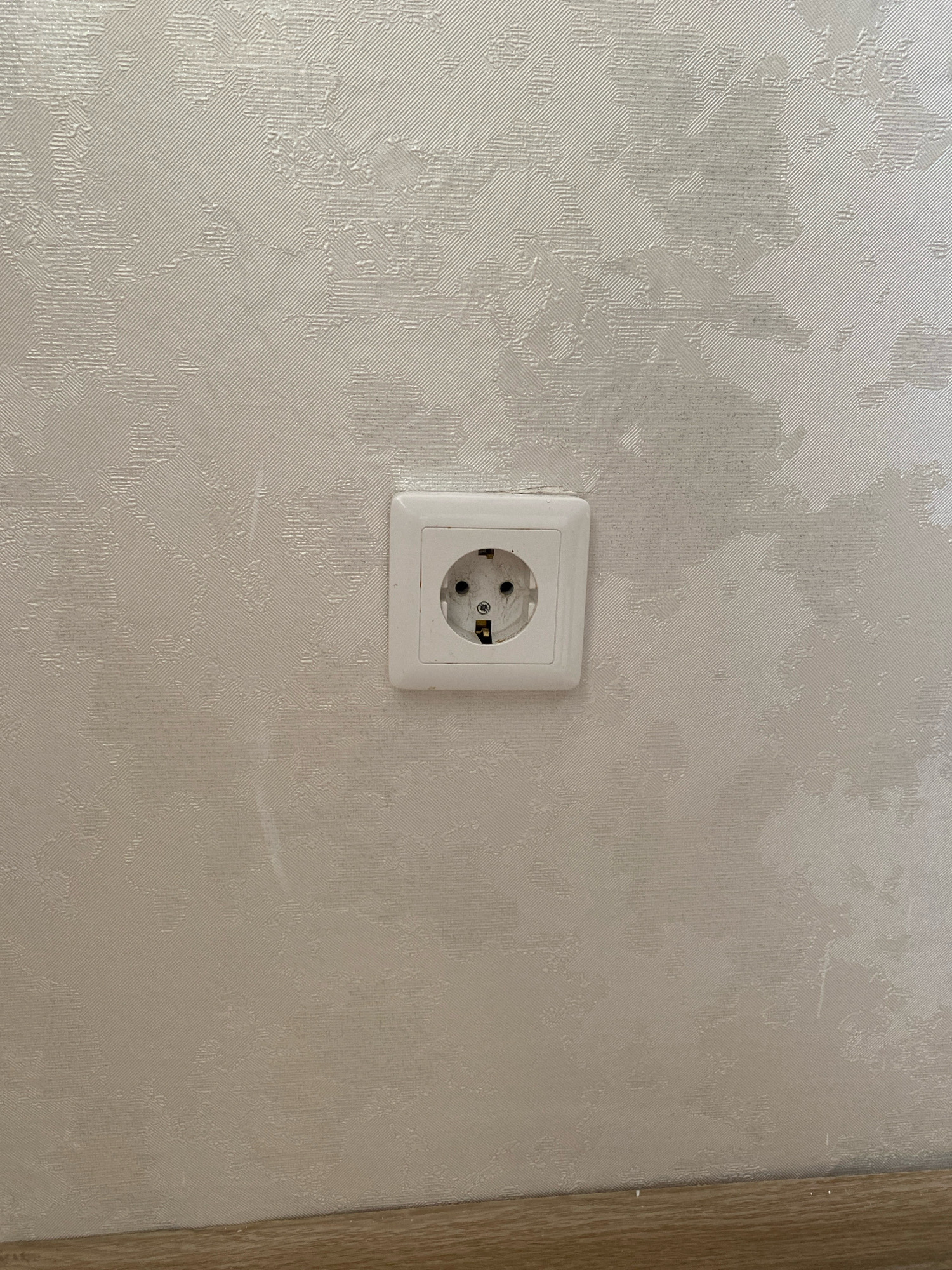 The image shows a white electrical outlet mounted on a textured, light-colored wall. The wall has a subtle, embossed pattern that appears to be abstract or floral. The outlet itself has a standard European design with two circular holes and a grounding contact. The outlet is positioned slightly off-center to the right. The bottom of the image shows a sliver of a light brown baseboard or trim. The scene appears to be indoors, possibly in a residential setting. There are no people or any discernable actions or interactions taking place. The location of Oyskhara, Russia, is not visually evident from the image itself.