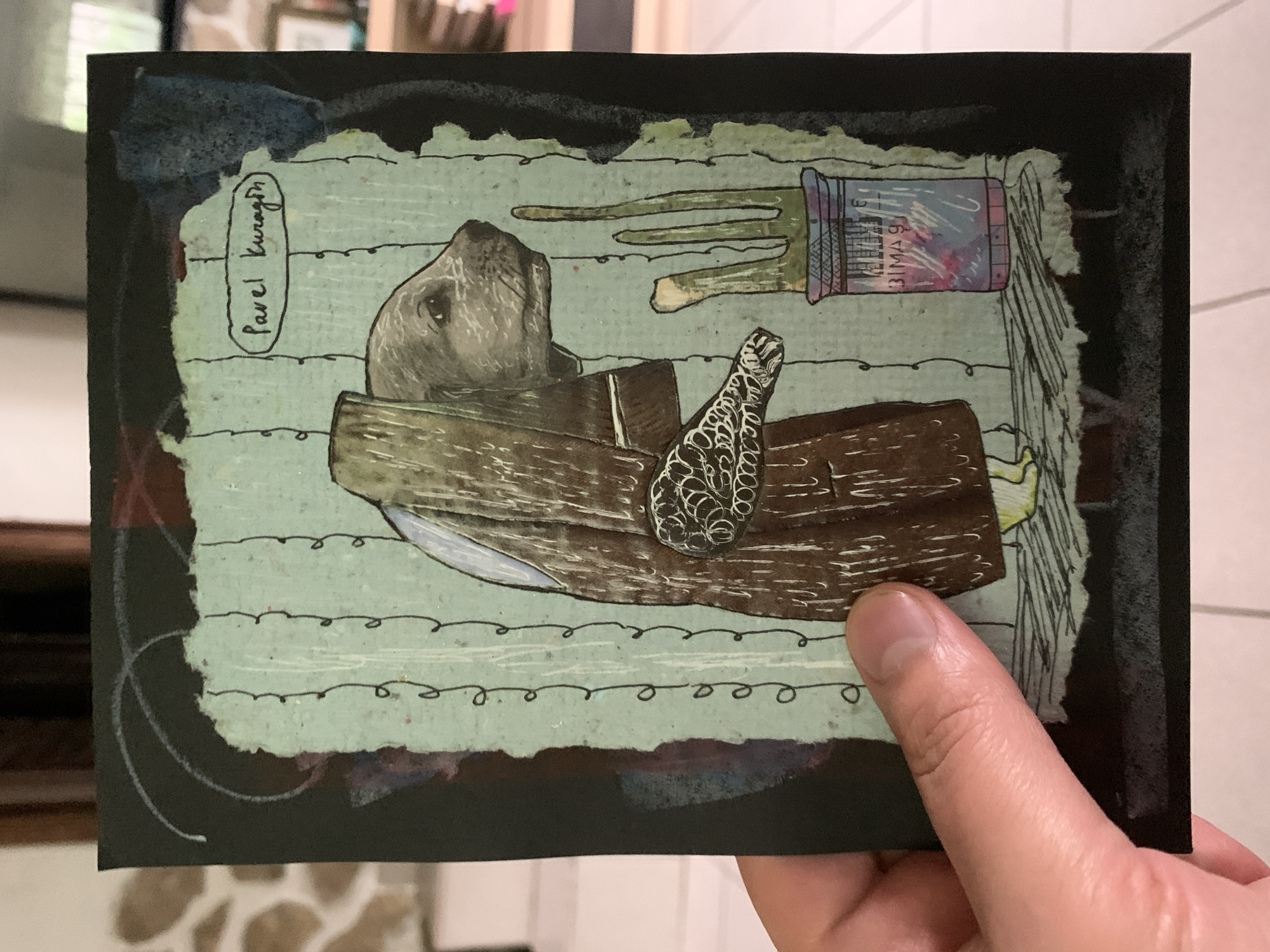 A postcard-sized artwork is held by a person's hand. The artwork features a stylized depiction of an animal, possibly a dog or a fox, wearing a brown garment and standing in front of a pale green background with vertical lines. To the right of the animal, a green, three-pronged object emerges from a cylindrical container. The container has a patterned design and text that appears to read "BIMAS". Above the animal's head, an oval shape encloses the handwritten name "Pavel Kuragin". The background of the artwork is black. The artwork itself is mounted on a larger black card. The scene appears to be indoors, with a blurred background that suggests a domestic or office environment. The location of Sergeevo, Russia, is not visually represented or implied by the artwork or its surroundings.