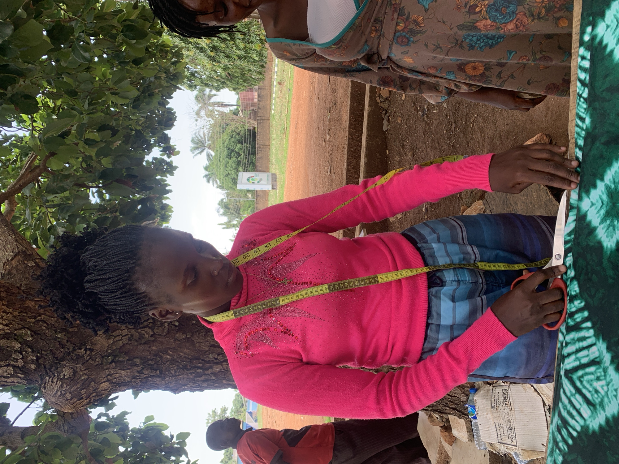 A woman in a bright pink sweater with rhinestone detailing and a measuring tape draped around her neck is cutting fabric with orange-handled scissors. She is wearing a blue and black striped garment and is positioned behind a piece of green tie-dye fabric. Another person, partially visible, is wearing a floral patterned garment. They appear to be outdoors, with a tree to the left, and a view of a grassy area with a sign and palm trees in the background. Debris, including a cardboard box, lies on the ground. The location is Maigizo, Nigeria.