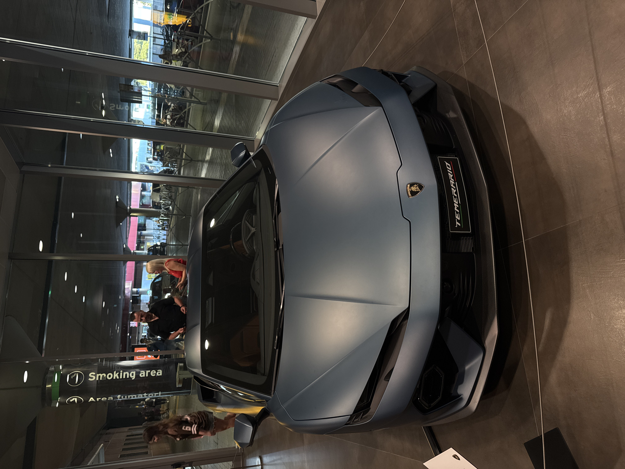 A matte blue Lamborghini sports car is displayed indoors, possibly in a showroom or at an event. The car has a prominent Lamborghini emblem on the hood and a "TEMERARIO" badge on the front grille, which displays the Italian flag colors. The vehicle's interior appears to be a tan leather.
Behind the car, large glass windows reflect an outdoor area with what appears to be a transportation hub, possibly an airport or train station, with people, seating areas, and vehicles visible. A sign with symbols of a cigarette and the words "Smoking area" and "Area fumatori" is visible to the left, suggesting a public space.
In the background, several individuals are present. To the left, a woman in a casual outfit walks away from the camera. To the right of the car, a man in a black shirt and a woman in a red top are interacting, possibly looking at the car or speaking with each other. The floor is tiled with a dark material. The overall lighting suggests it is either nighttime or an indoor space with artificial lighting. The presence of the "Smoking area" sign and the architectural style of the background point towards a public transit terminal. Given the context of the "TEMERARIO" model, this might be a special exhibition. The city of Bologna, Italy is known for its automotive industry and historical sites, but this scene primarily depicts a modern commercial or transit environment.