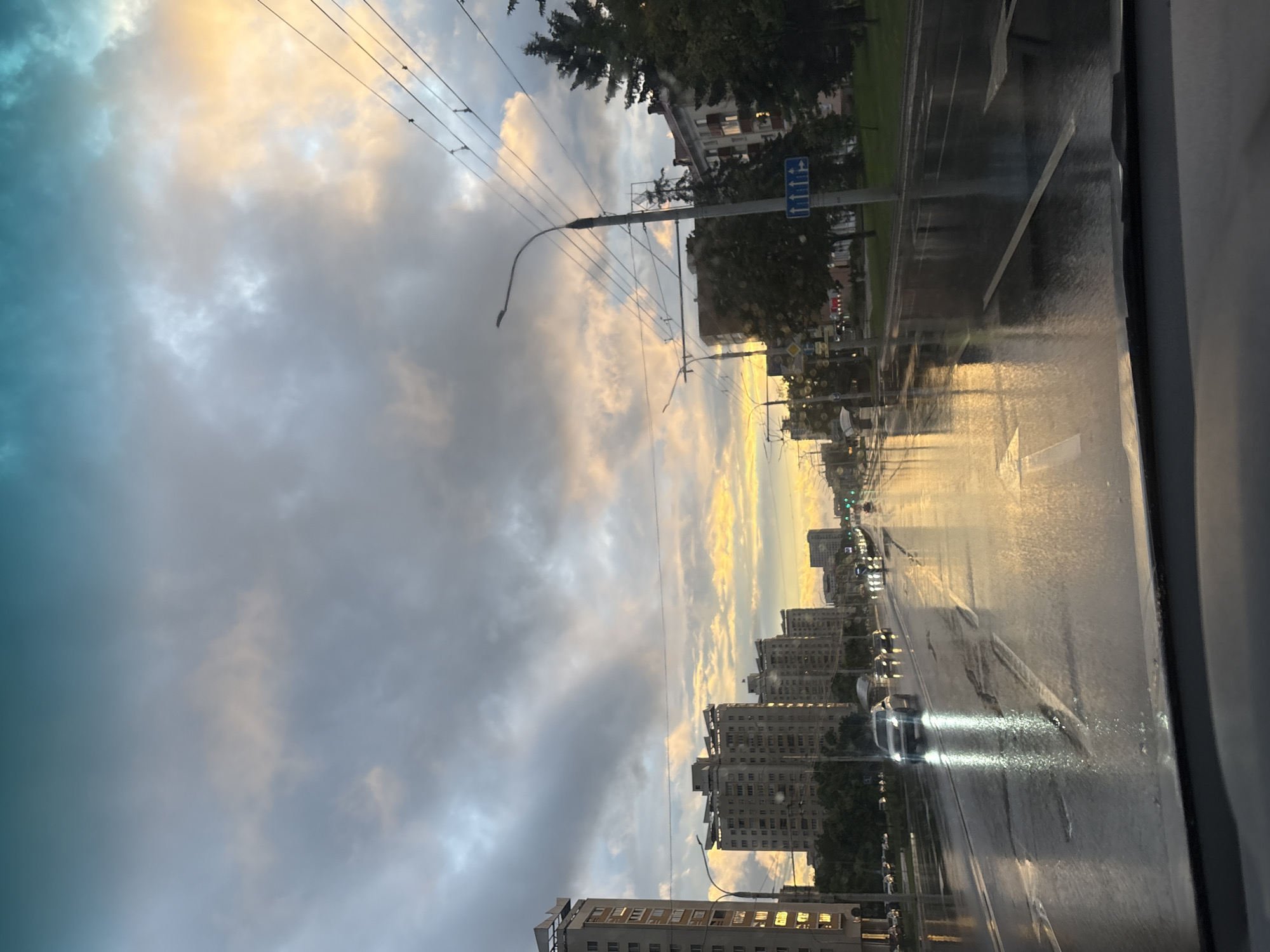 The image depicts a street scene in Minsk, Belarus, during a cloudy sunset. The road is wet, reflecting the sky and the headlights of approaching cars, suggesting recent rainfall. Tall apartment buildings line both sides of the wide, multi-lane road. Overhead wires, likely for public transportation, crisscross the sky. The sky is a dramatic mix of grays, blues, and warm oranges and yellows, indicative of the setting sun breaking through the clouds. Traffic lights are visible in the distance, and streetlights are illuminated. There are no visible people in the image, but several cars are on the road, some with their headlights on. The overall atmosphere is one of a wet, urban evening.