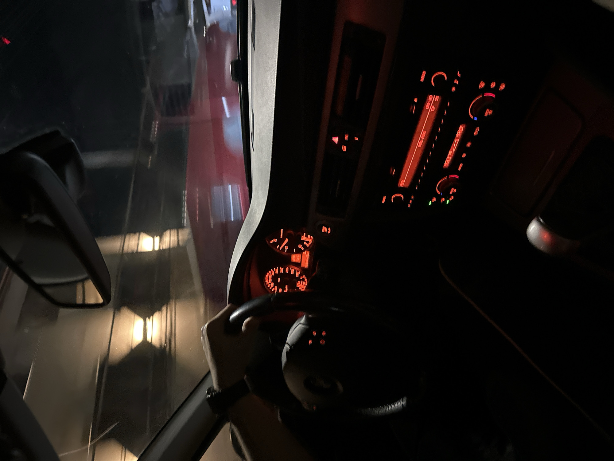 A view from inside a car at night. A hand grips the steering wheel. The dashboard is visible, illuminated with red lights. Outside the window, a blurred view of buildings and lights suggests an urban environment. The image does not provide enough information to confirm the location is Polena, Bulgaria.
