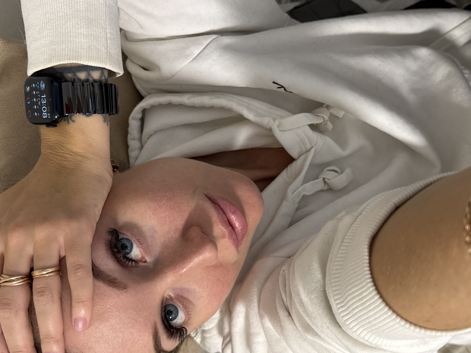 The image captures a close-up, slightly tilted view of a person, likely female, lying down and looking towards the camera. The subject has fair skin, blue eyes with prominent eyelash extensions, and well-groomed eyebrows. A small nose stud is visible on her nose, and her lips appear lightly glossed. She is wearing a white or off-white hooded sweatshirt, with its hood and drawstrings visible around her neck and shoulders.
On her left wrist, she wears a black Apple Watch with a sleek, black link band. The watch display clearly shows the time as "13:08" (1:08 PM) and the date as "ПТ 24" (Friday 24th), along with small icons indicating weather and activity rings. A tattoo is partially visible underneath the watch band on her forearm. Her right hand, adorned with multiple gold-toned rings on her fingers, rests near her face, with the fingertips gently touching her temple.
The background consists of soft, neutral-toned fabric, suggesting the person is lying on a bed or sofa. A small, stylized dark script, possibly a logo or initial, is subtly visible on the white hoodie near the neckline. The overall mood is relaxed and serene. The location context for this image is Chaikovsky, Russia.