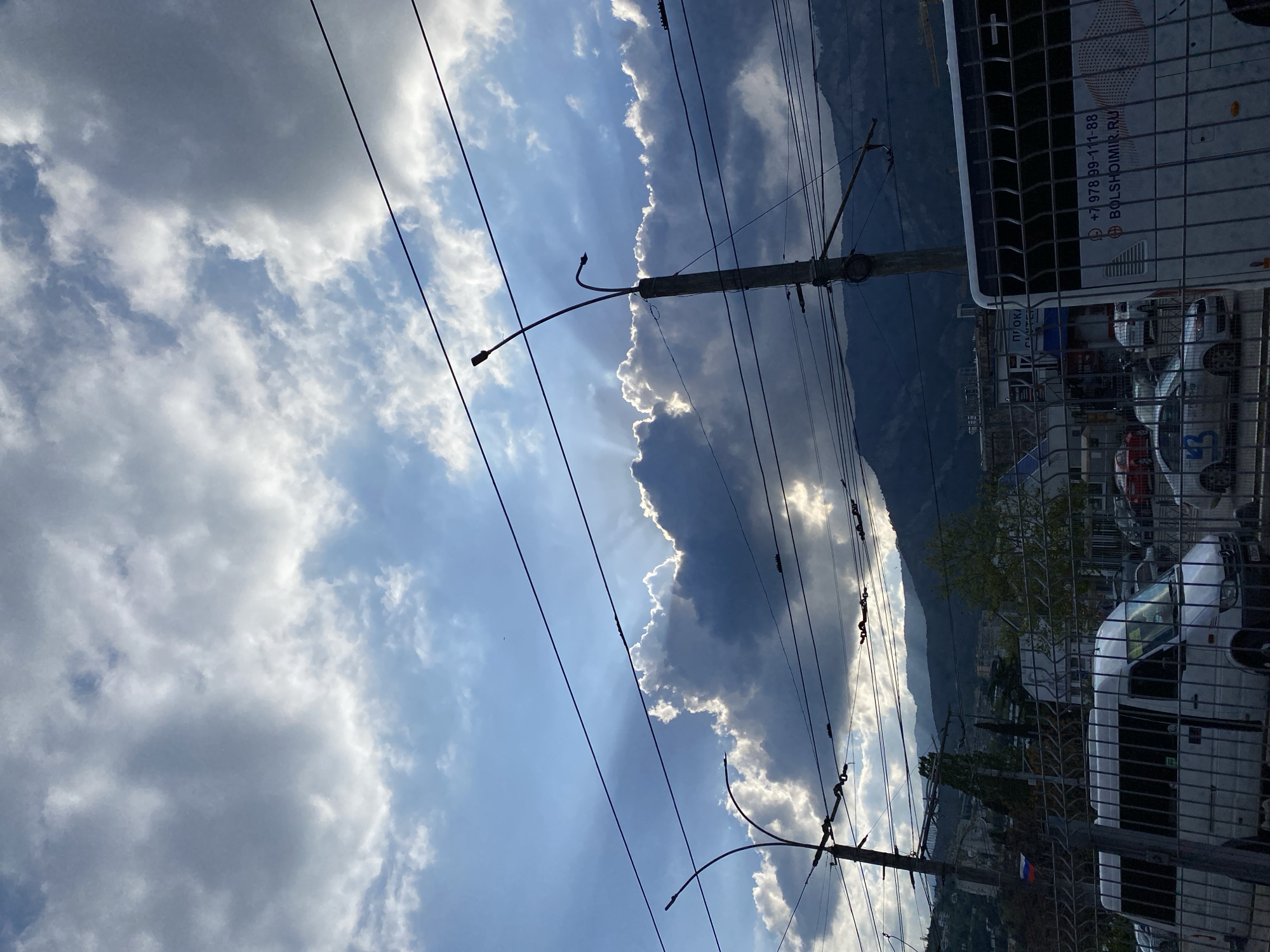 The image shows a view in the city of Ялта, Ukraine. The sky is visible with a mix of light and dark clouds, with sun rays filtering through. There are power lines running across the scene, supported by utility poles. In the background, mountains are partially obscured by clouds. A white van is visible on the right side of the frame, partially obscured by a metal fence. Other vehicles and buildings can be seen in the distance.