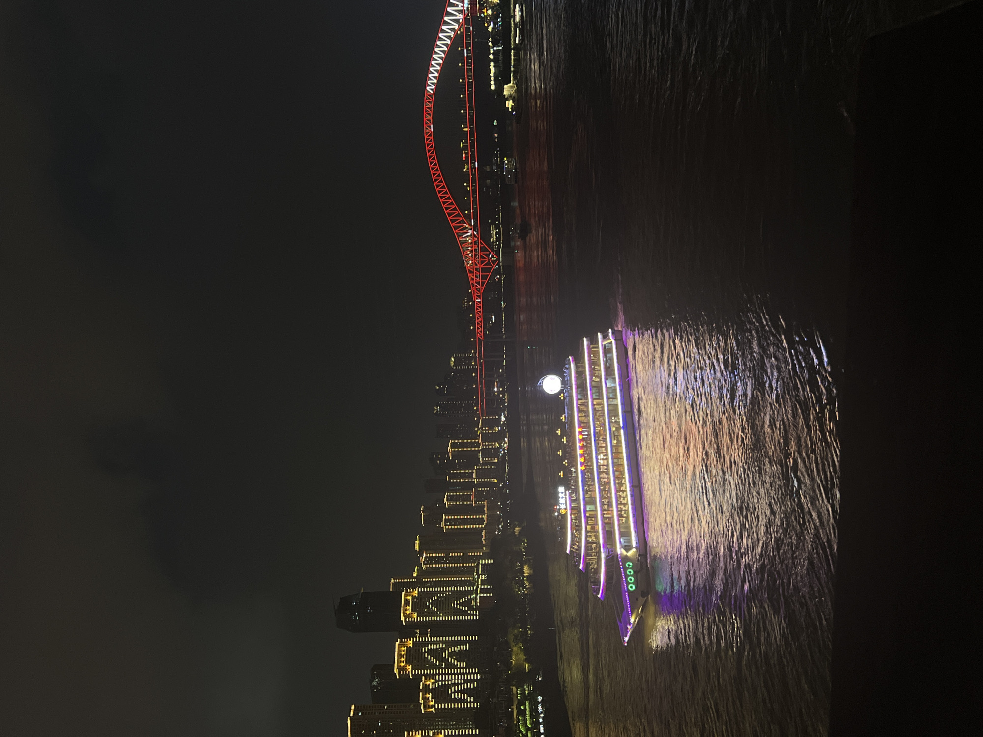 The image captures a nighttime view of the Yuzhong District in China, likely from a vantage point overlooking the Yangtze River. A large, illuminated cruise ship, adorned with purple and white lights, sails on the dark water in the foreground. The ship's lights reflect on the river's surface, creating a vibrant glow. In the background, a cityscape of tall buildings, brightly lit with yellow and white lights, stretches across the skyline. A distinctive red-lit bridge with an arching structure spans the river, adding a striking architectural element to the scene. The overall ambiance is one of a bustling, modern city illuminated against the dark night sky.