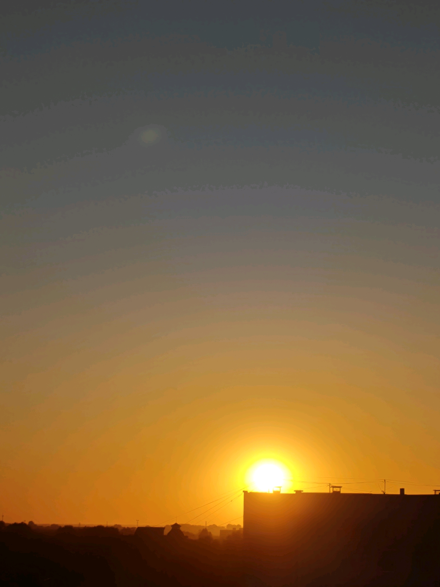 The image captures a sunset over Rubtsovsk, Russia. The sun, a brilliant orange orb, is partially obscured by the silhouette of a flat-roofed building, likely an apartment complex or industrial structure. The sky transitions from a deep blue at the top to a warm gradient of yellow and orange near the horizon, indicating the sun is low in the sky. The lower portion of the image shows a dark, silhouetted landscape with the faint outlines of other buildings and structures, suggesting an urban or semi-urban environment. No people are visible in the frame. The overall scene is one of a calm, late evening in a Russian city.