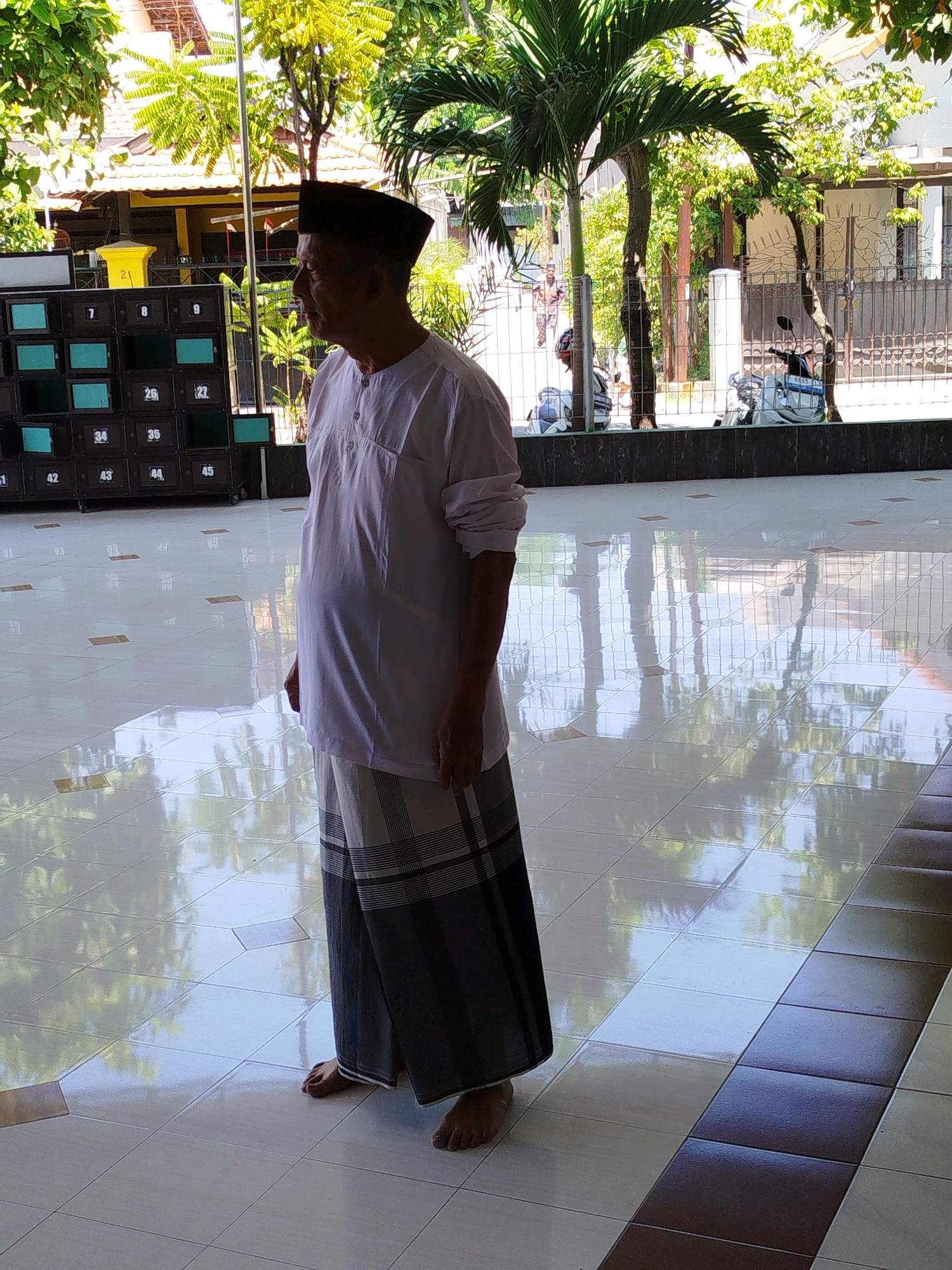 An elderly East Asian man wearing a white long-sleeved shirt, a black peci (cap), and a sarong stands barefoot on a tiled floor, looking to the left. He is in a covered outdoor area with a tiled floor that reflects the surrounding environment. In the background, there are lockers with numbered compartments, palm trees, and a building with a yellow facade. Beyond that, a fence separates the area from a street where two motorcycles are parked and a person is walking. The scene appears to be a public space, possibly a mosque or religious center, given the attire of the man and the presence of lockers which could be for shoes or personal belongings. The lighting suggests it is daytime. The location is identified as Surabaya, Indonesia.