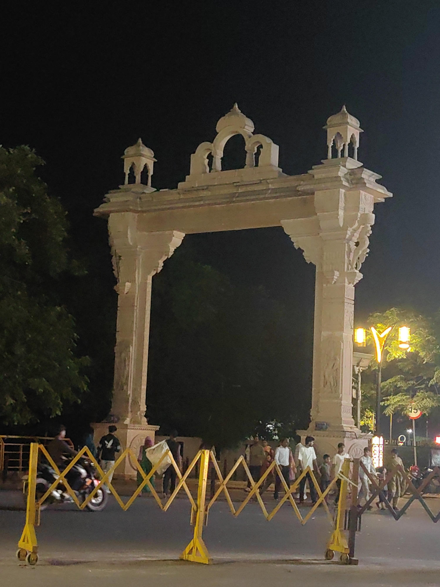 This image captures a night scene at the gate of Gorakhnath Temple in Gorakhpur, India. The prominent structure is a large, ornately carved sandstone archway with multiple domes and decorative elements. In the foreground, a yellow, expandable barricade with wheels is set up across the road. To the left of the archway, a person is riding a motorcycle. Several other individuals are visible walking near the archway and across the road, some carrying bags. Streetlights illuminate the scene, casting a warm glow on the architecture and the surrounding trees. The overall atmosphere suggests a busy evening at a significant religious and cultural landmark.