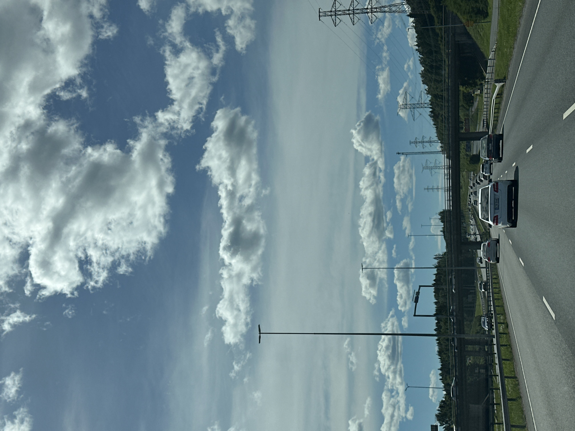 The image captures a view from a moving vehicle on a multi-lane highway under a partly cloudy sky. The highway has at least three lanes visible in the foreground, with several cars driving in the same direction. An overpass stretches across the highway in the mid-ground, with trees and more cars visible on it. In the background, more trees and power lines with tall pylons are present. Tall streetlights are also visible along the side of the highway. The sky is a vibrant blue with numerous fluffy white clouds. The overall scene depicts a typical highway environment on a bright day.