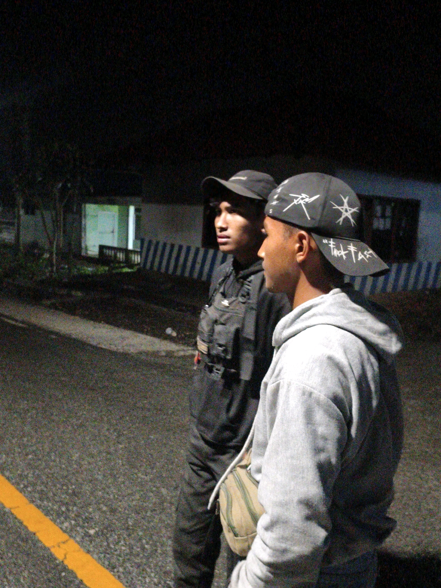 Two young men are standing on an asphalt road at night. The man on the left is wearing a black baseball cap, a black tactical vest over a black shirt, and dark pants. The man on the right is wearing a black baseball cap with white star-like graphics and text, a grey hooded sweatshirt, and dark pants. He has a canvas messenger bag slung across his body. In the background, there is a building with striped blue and white railings and some trees. A yellow painted line is visible on the left side of the road. The scene is dimly lit, suggesting an outdoor urban or semi-urban environment.