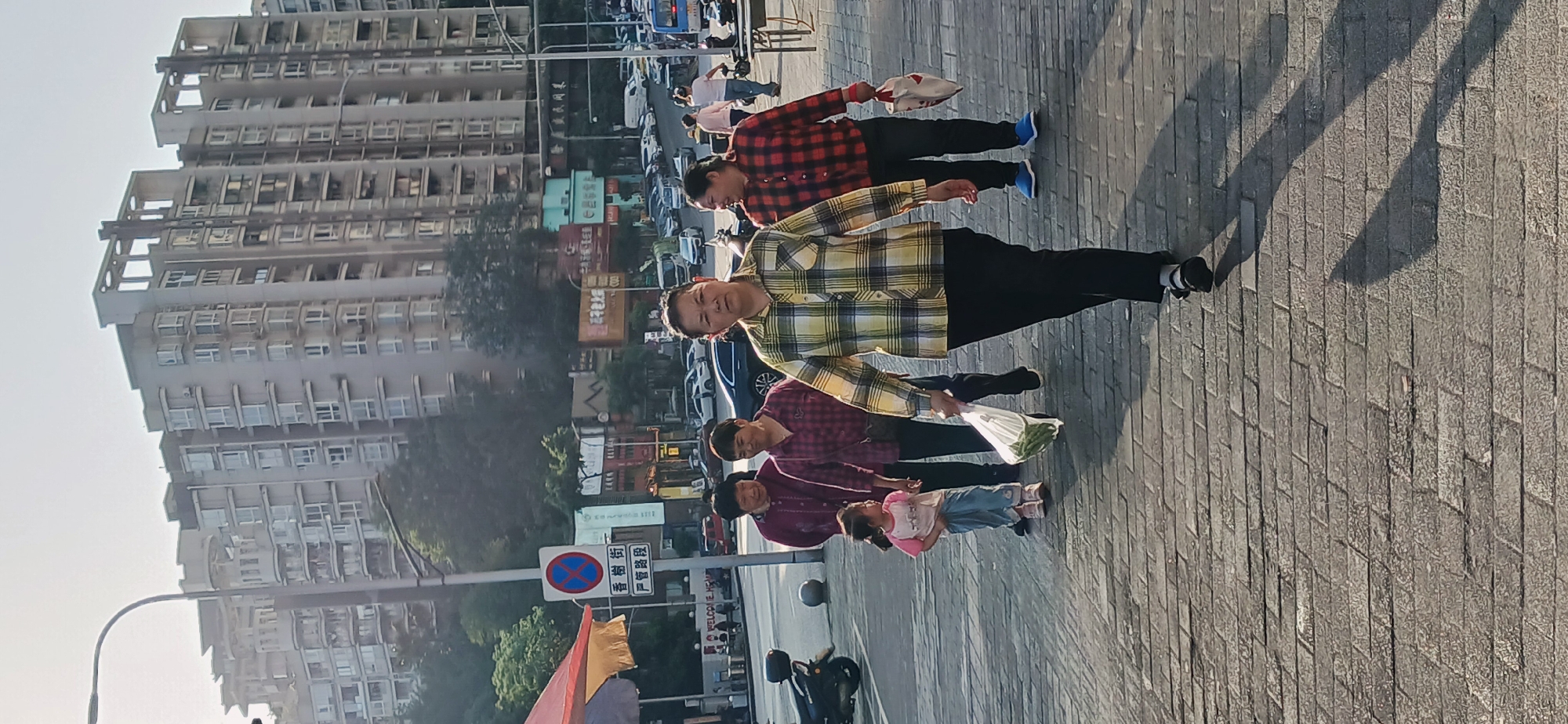 A group of five people, including a child, walks along a brick-paved sidewalk in an urban area. The backdrop consists of tall apartment buildings and a busy street with cars and pedestrians. The group includes adults wearing plaid shirts, one of whom is holding a white plastic bag filled with vegetables. A "No Parking" sign is visible near the group. The location is identified as Jiulongpo District, China, suggested by the language on the sign and architectural styles. A motorcycle is parked close by.