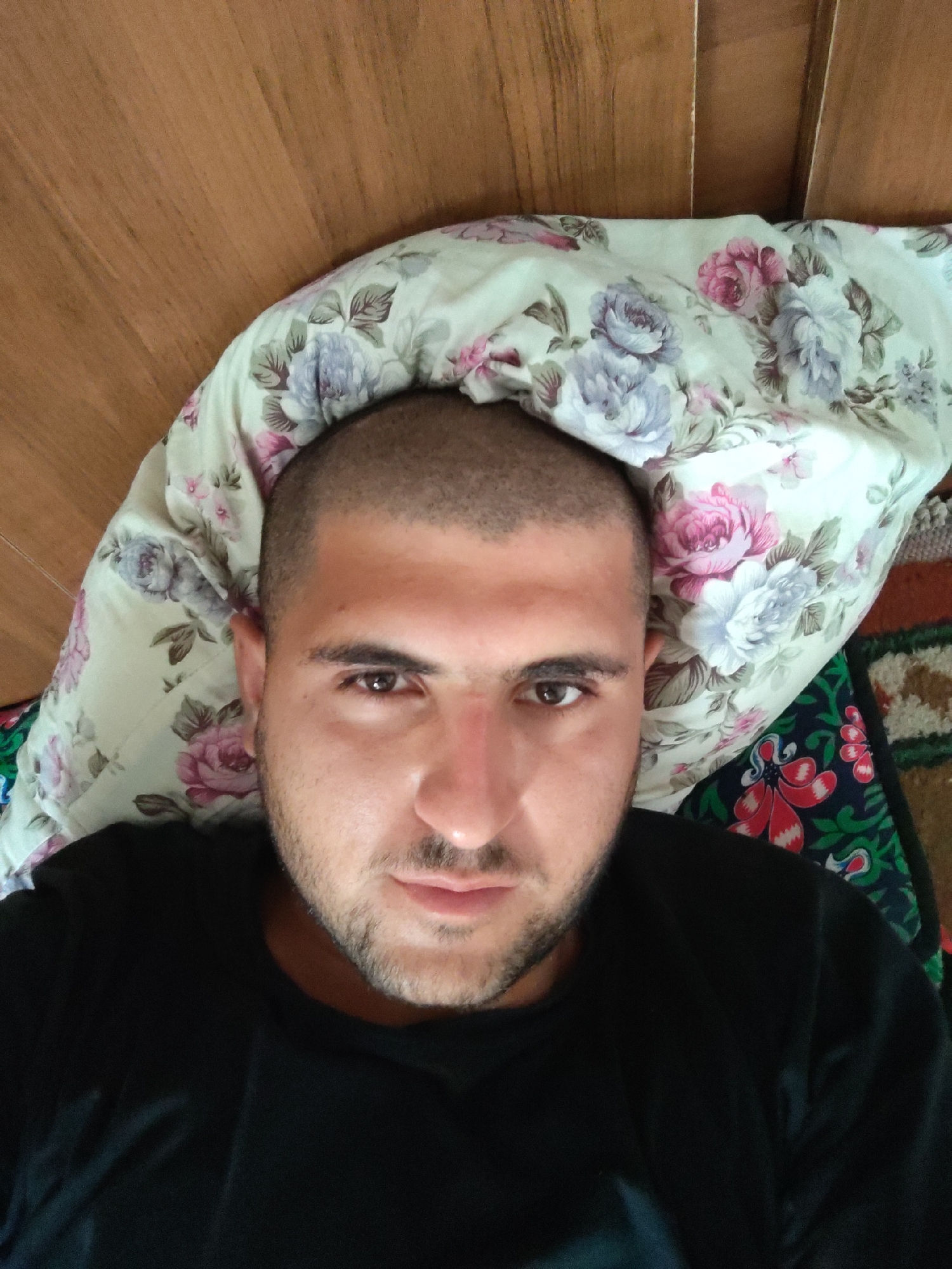 A close-up, overhead, selfie-style shot shows a man with a shaved head and short beard lying on his back. He is wearing a black t-shirt and is positioned on a floral-patterned pillow. The pillow is on a bed with a wooden headboard visible in the background. To the right of the pillow, a colorful patterned blanket is partially visible. The lighting suggests it is daytime, likely indoors. The image is devoid of any specific landmarks or contextual elements that would indicate its location within the city of Khujand, Tajikistan.