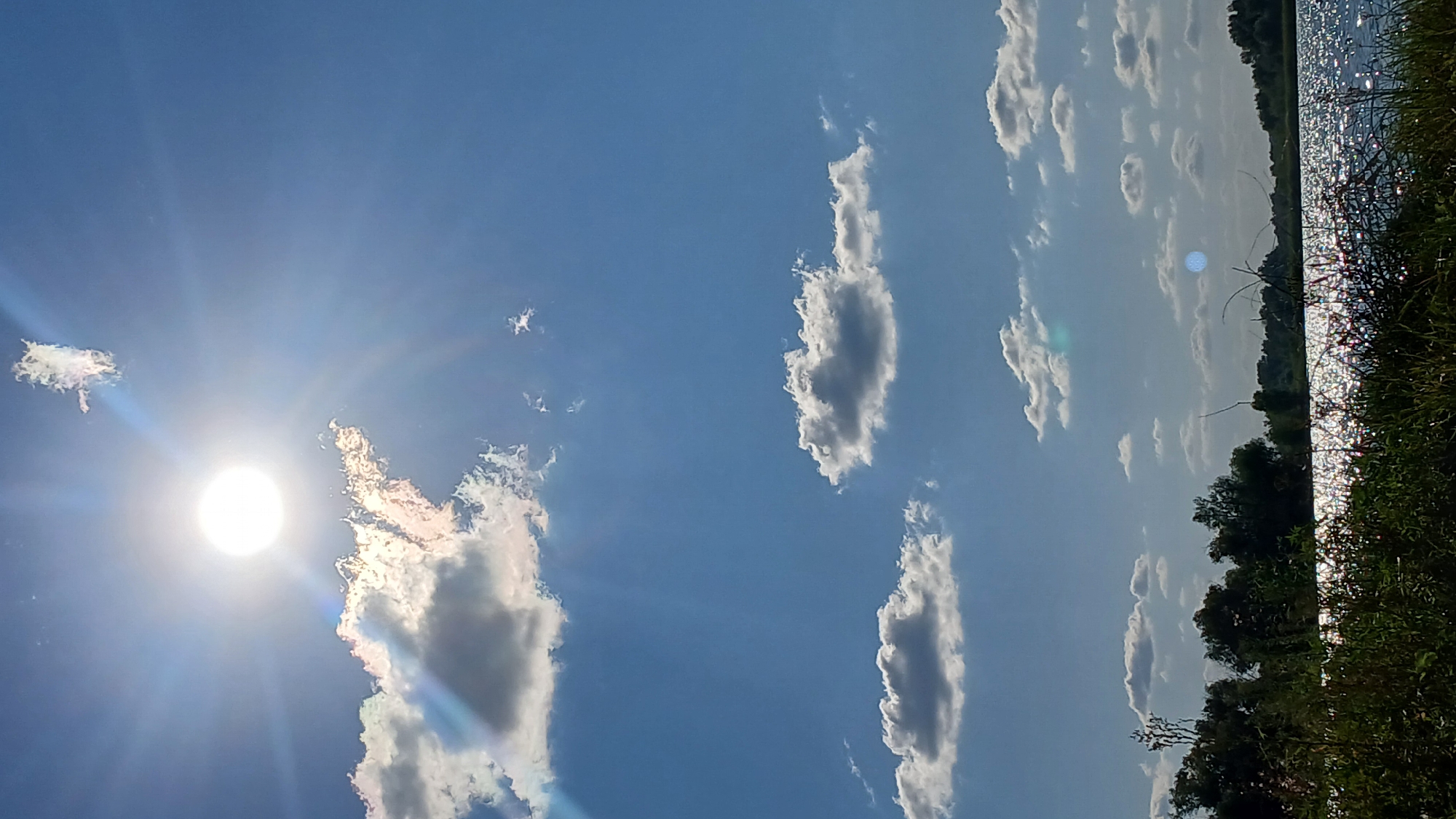 The image depicts a bright, sunny day with a clear blue sky dotted with white, puffy clouds. The sun is a prominent feature, emitting strong rays that create a lens flare effect. In the foreground, there is a body of water, likely a lake or river, reflecting the sunlight and creating a shimmering effect on its surface. The shoreline is lined with lush green vegetation, including trees and tall grass. The overall scene suggests a natural landscape in a rural or suburban setting. The provided location of Jelaevo, Kazakhstan, aligns with this description of a natural environment.