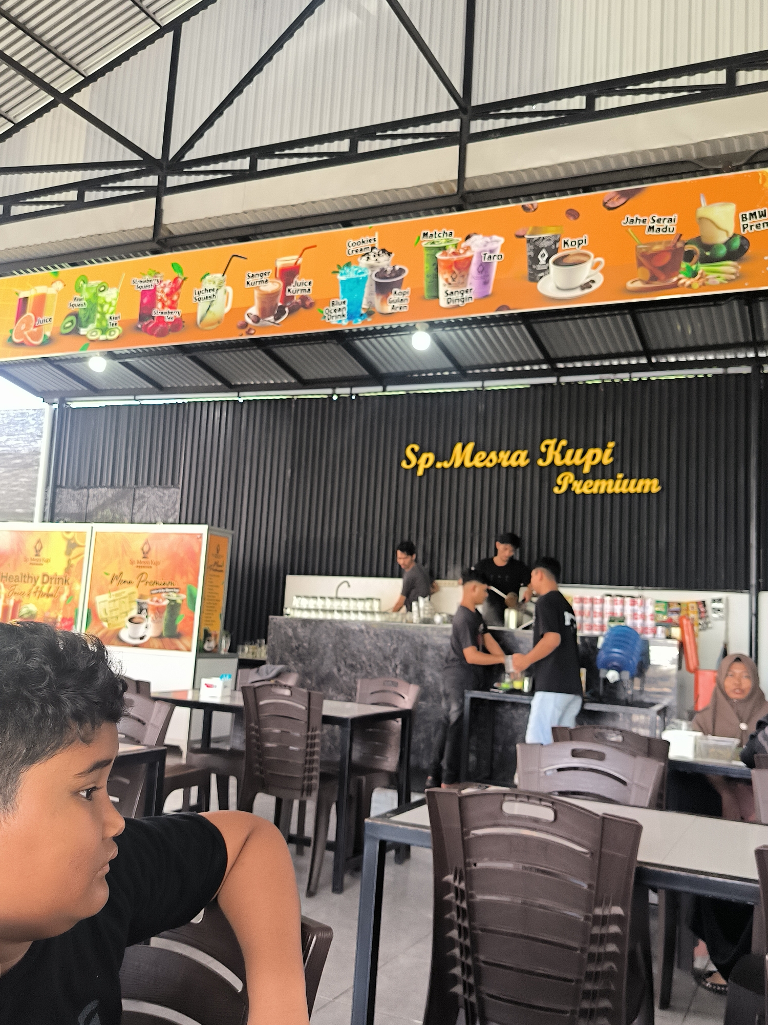 This image depicts the interior of "Sp. Mesra Kupi Premium," a cafe or restaurant, likely located in Banda Aceh, Indonesia. In the foreground, a young boy with dark, curly hair is seated at a table, looking contemplatively to his left. The cafe features multiple dark-colored tables and brown plastic chairs for patrons.
Behind the seating area, a long counter serves as the main preparation and service point, where several male staff members are busy. The wall behind the counter is dark with vertical ribbed panels, featuring a prominent yellow sign with the cafe's name, "Sp. Mesra Kupi Premium." Above the counter, a large orange menu board showcases a variety of drinks with illustrative images. Visible drink names include "Juice," "Kiwi Squash," "Strawberry Squash," "Lychee Squash," "Sanger Kurma," "Juice Kurma," "Cookies Cream," "Matcha," "Taro," "Blue Ocean Drink," "Kopi Gulan Aren," "Kopi," and "Jahe Serai Madu." Two smaller display boards are also visible, one labeled "Healthy Drink Juice & Herbal" and another "Menu Premium."
The cafe has an open, industrial-style ceiling with exposed metal roof trusses and a corrugated metal roof. Natural light suggests it is daytime. In the far right background, a woman wearing a hijab is seated at a table. The overall scene portrays a casual and busy cafe environment.