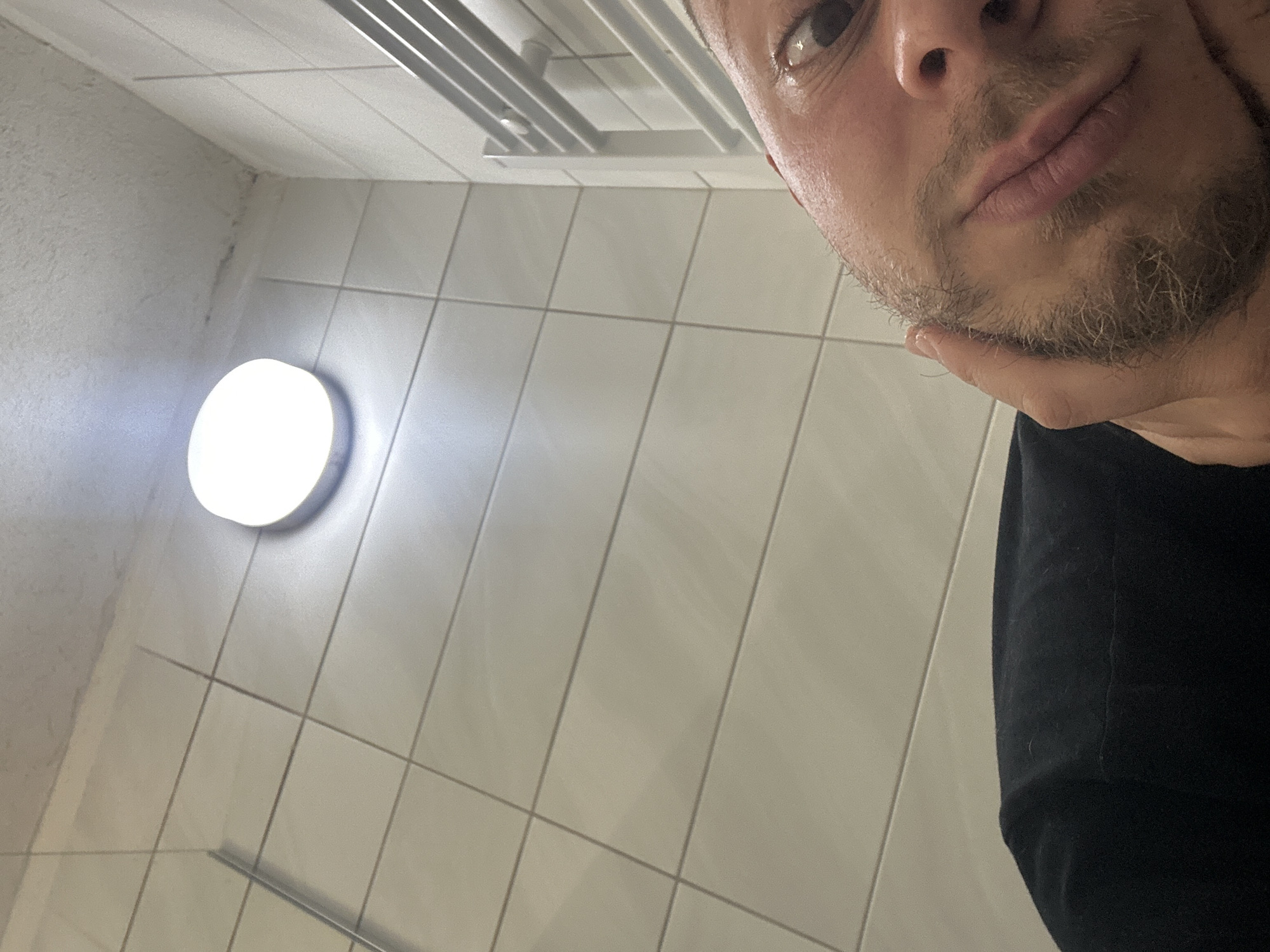 The image presents a close-up view of a person's face against a backdrop of a tiled wall and ceiling fixtures. The person has fair skin, light-colored hair, and a trimmed beard. They are wearing a black garment. Their hand is positioned near their chin.
The environment suggests a utilitarian space, possibly a bathroom or restroom, indicated by the white tiles and the circular ceiling light. A metal fixture, potentially a towel rack or ventilation system, is visible near the ceiling.
The location is in the city of Unknown, Unknown.