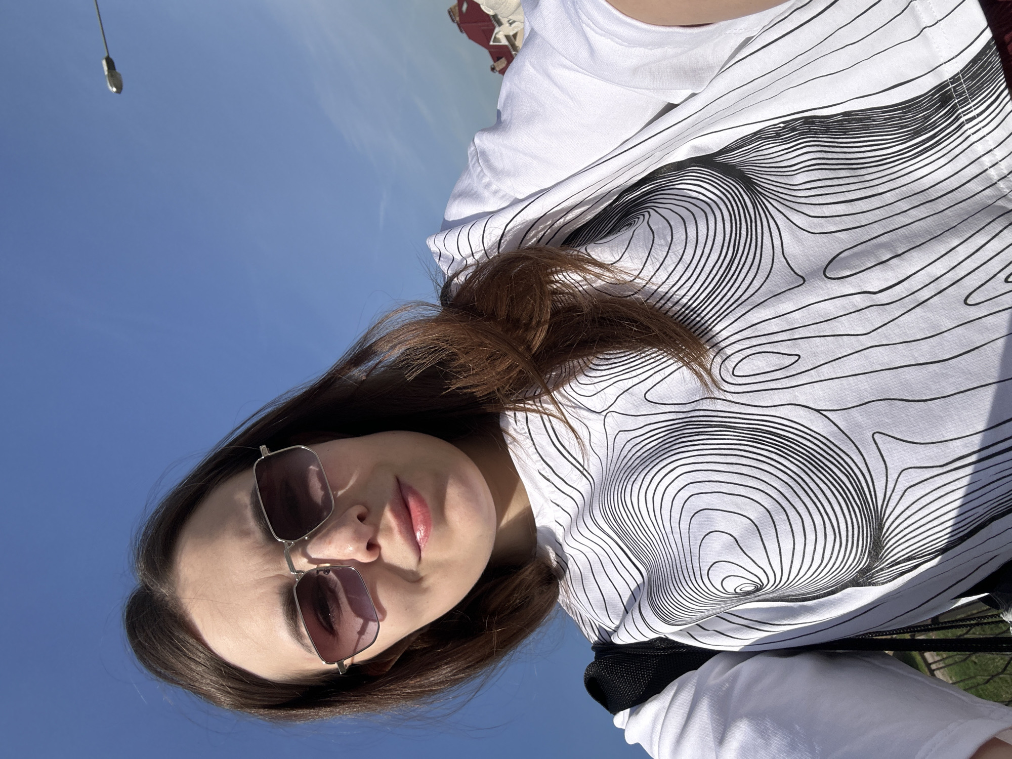 A selfie taken from a low angle shows a young woman wearing sunglasses and a white t-shirt with a black geometric pattern against a bright blue sky. The photo was taken during the day. A blurred structure is in the upper left, and vegetation is visible in the lower right. Based on this information, the image can be identified as likely having been taken in Perm, Russia.