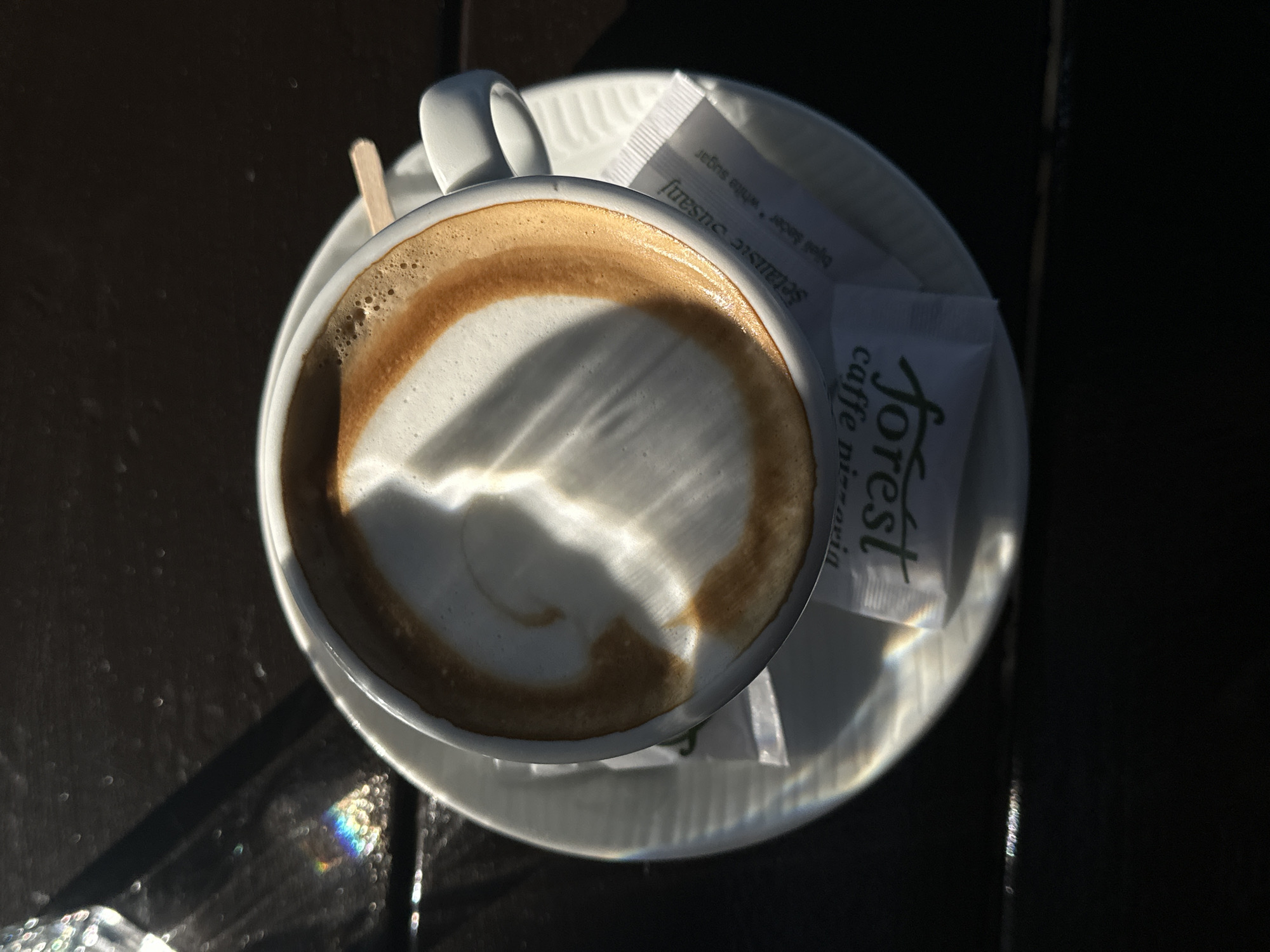 The image shows a top-down view of a cappuccino on a white saucer, placed on a dark surface that appears to be a table. Next to the cup are two packets of sugar. The branding on one sugar packet reads "Forest caffe pizzeria", suggesting the coffee was obtained from a Forest caffe pizzeria establishment, which are present in Šušanj, Montenegro. Light reflects off the foam of the cappuccino. A wooden stir stick is also visible. The surface is likely outdoors, with varying light and shadow.