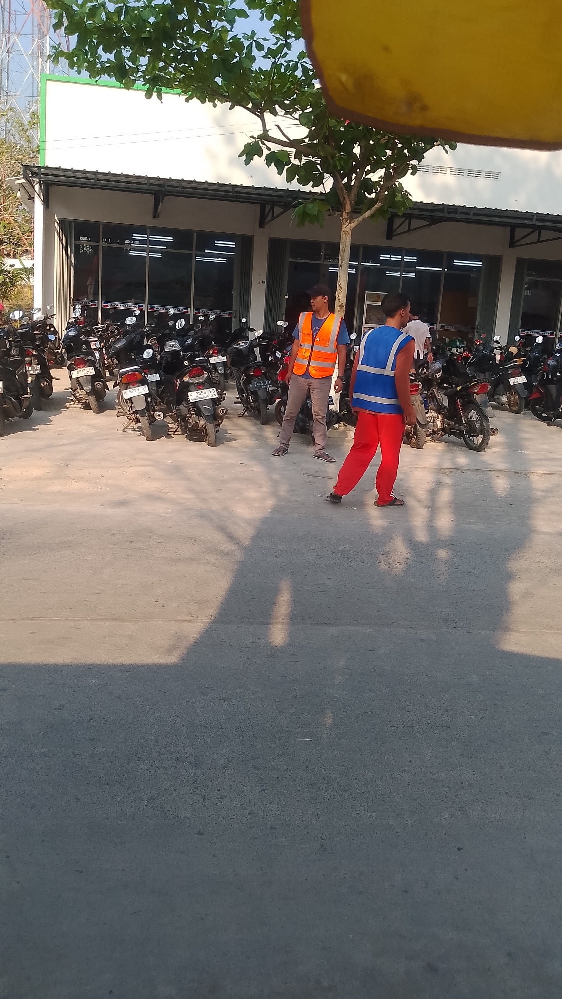 In Karanglangu, Indonesia, a paved outdoor parking area is situated in front of a modern commercial building with a green trim and a metal overhang. A dense collection of parked motorcycles fills the foreground and midground, with their handlebars and seats visible. Two men are present in the scene. One man, wearing a blue shirt and grey pants, stands facing left and is mostly obscured by a bright yellow, blurry object in the upper right corner of the image. The second man, wearing an orange safety vest over a blue shirt and grey pants, stands to the left of the first man, facing the camera. A third individual, wearing a blue vest and red pants, is seen walking away from the camera toward the right side of the image, with their back turned. The sun casts long shadows across the pavement, indicating it is likely late afternoon. The building facade has multiple large windows with internal lighting visible. A large tree with green leaves grows in front of the building. The sky is clear and blue.