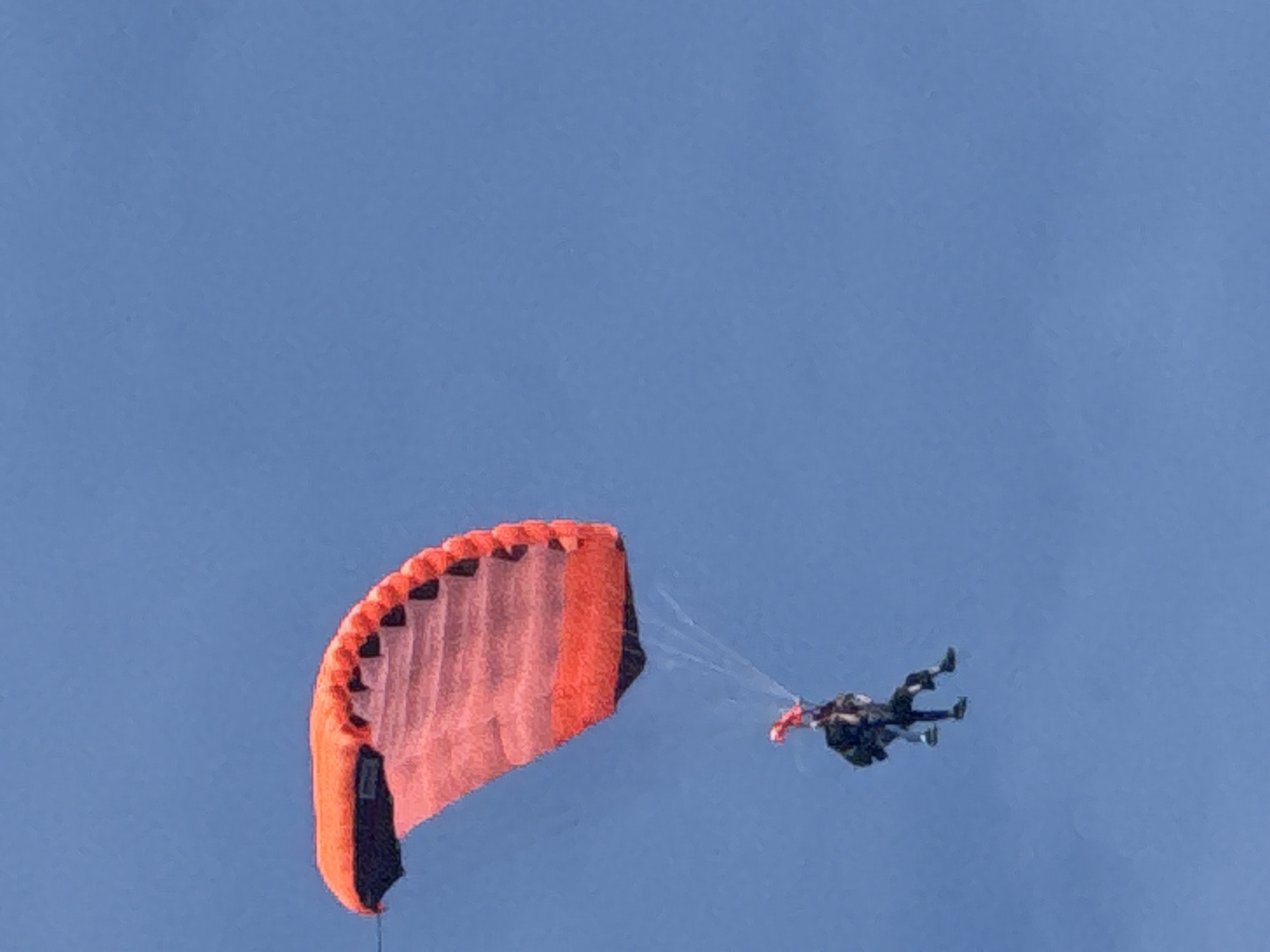 A tandem skydiving pair descends against a clear, uniformly bright blue sky in Unknown, Unknown. The primary object is a large, rectangular ram-air parachute canopy, predominantly vibrant orange and red with black detailing along its edges and trailing surface. White suspension lines extend downwards from the parachute. Below, two individuals, clad in dark jumpsuits, are visibly strapped together in a tandem harness configuration, suspended in a seated posture. One individual is positioned slightly above and behind the other. The scene depicts an outdoor aerial activity under daylight conditions, with no other environmental features or objects present.