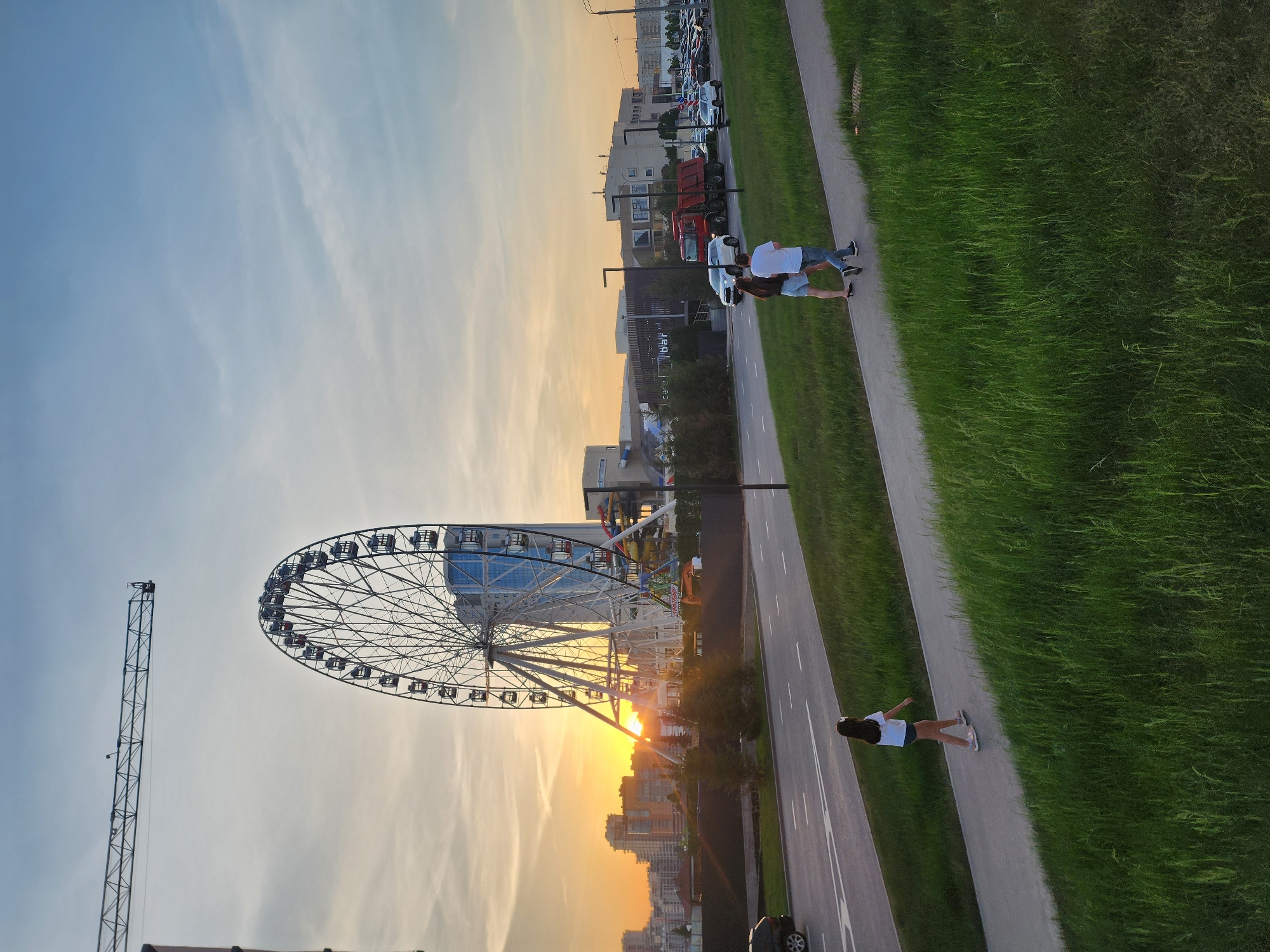 The image, taken in Kazan, Russia, features a Ferris wheel in the foreground, silhouetted against a setting sun. A tall crane stands adjacent to the Ferris wheel. In the background, city buildings are visible. A road runs parallel to a grassy area. On a pathway between the road and grass, a woman is lying down with her arms outstretched, while two people are walking in the opposite direction. A car is parked along the road.