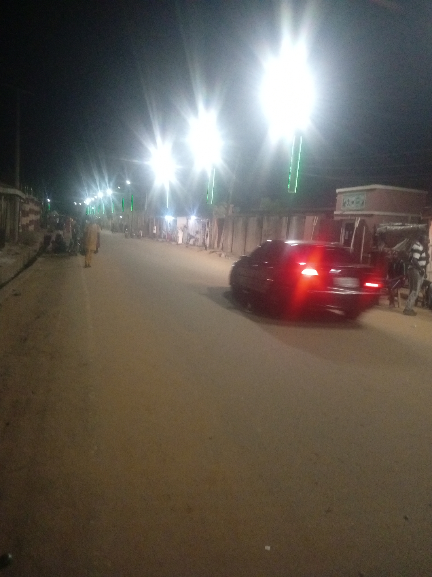 The image depicts a street scene at night in Tundun Wada West, Nigeria, illuminated by streetlights and the taillights of a dark-colored car. The road surface is paved and appears to be made of asphalt or a similar material. The car, a sedan with its brake lights illuminated, is in motion on the right side of the road. Along the sides of the street, there are buildings and structures, some of which appear to be shops or homes. Several people are visible, including individuals standing on the side of the road and a person standing near the car. There are also motorcycles parked along the side of the road. The overall impression is that of a moderately busy street at night, with artificial lighting providing visibility.