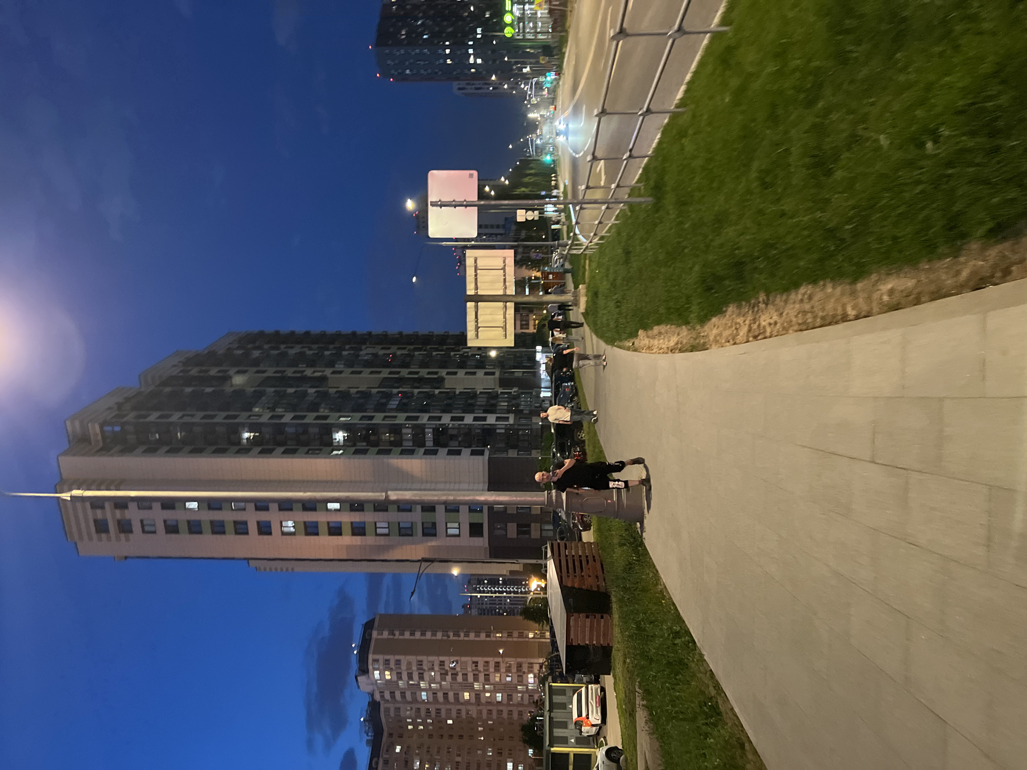 The image shows an urban scene at dusk in Vnukovo, Russia. A paved sidewalk runs along a grassy area adjacent to a road. On the sidewalk, there are people walking, and one person appears to be standing next to a trash can. Several multi-story residential buildings dominate the background. Streetlights are visible, and a few cars are on the road. The sky is a deep blue. Various road signs and barriers line the street.