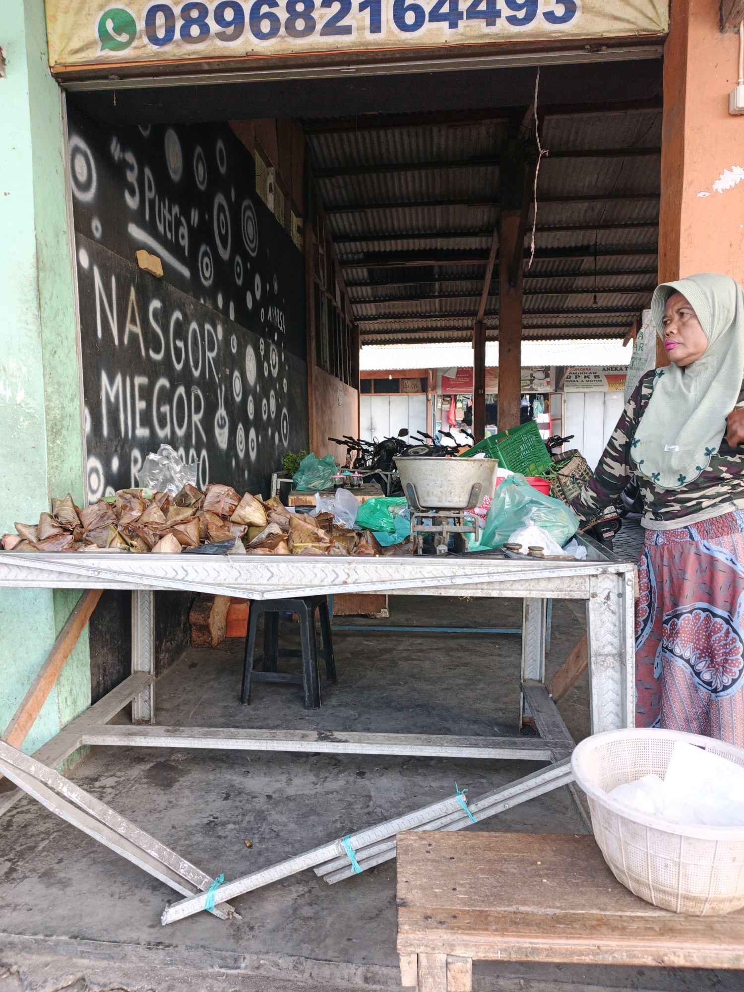 The image depicts an outdoor market stall in Kludan, Indonesia. A woman wearing a green hijab and a camouflage patterned shirt stands to the right, looking towards the left. In front of her, a metal table is laden with a large pile of food items wrapped in banana leaves, triangular in shape. To the left of the table, a black plastic stool is visible. Behind the table, a scale with a metal bowl sits on the counter. The background shows a corrugated metal roof supported by wooden beams, suggesting a covered market area. Graffiti art adorns the wall behind the stall, with the words "NASGOR MIEGOR" clearly visible, indicating food offerings. A mobile phone number, "089682164495", is displayed on a banner above the stall. The ground is a concrete surface.