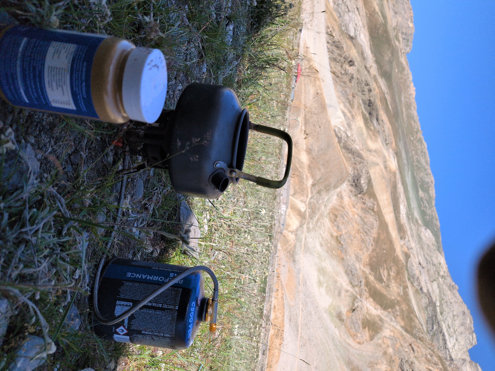 The image captures an outdoor scene with a mountainous, arid landscape in the background under a clear blue sky. In the foreground, on uneven ground covered with dry grass and rocks, are camping equipment. A dark metal camping kettle sits on a stove. To its right is a blue and black gas canister with a grey hose. In front of the kettle, a jar with a white lid is visible, with a label partially obscured. A small red building is faintly visible in the middle ground of the landscape. The location is identified as Matayeen, India. There are no people present in the image.