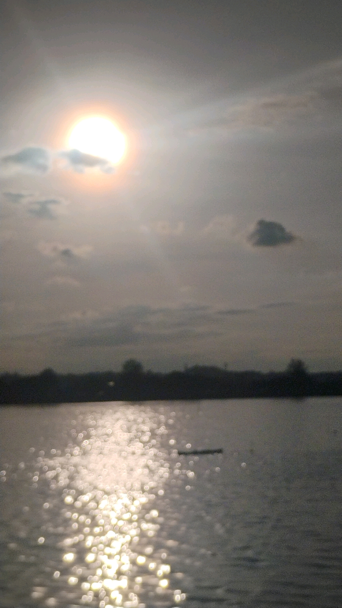 The image depicts a waterfront scene at sunset or sunrise, likely in Lhokseumawe, Indonesia. The sun, partially obscured by clouds, casts a bright glare on the water, creating a bokeh effect. A dark, indistinct landmass with silhouetted trees forms the horizon. The water itself is calm, with a small, dark object resembling a narrow boat or raft floating on the surface. The overall atmosphere is serene and tranquil.