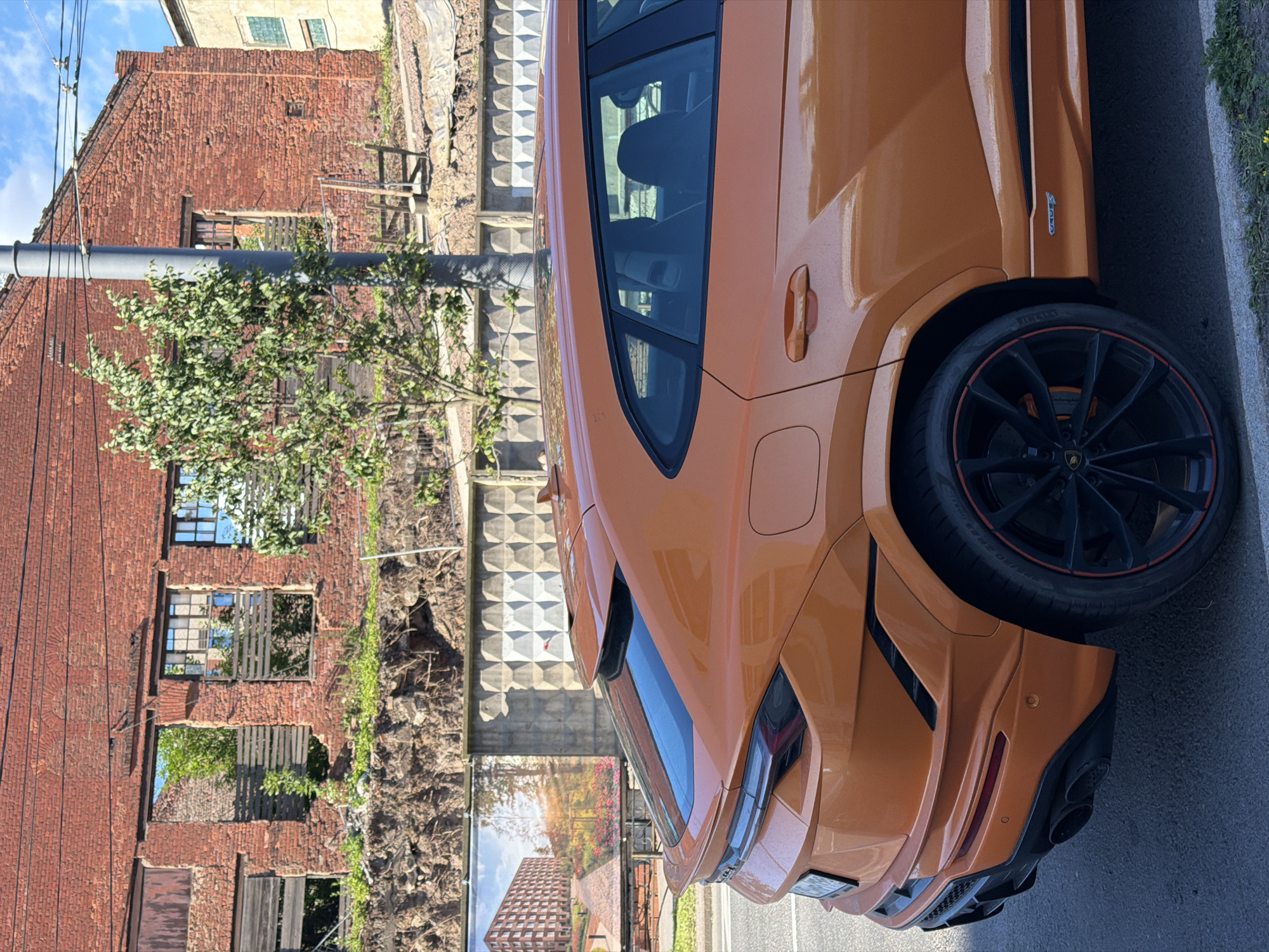 An orange Lamborghini Urus is parked on an asphalt road in front of a derelict red brick building in Saint Petersburg, Russia. The building has broken windows and appears to be under construction or demolition. A billboard displaying an architectural rendering of a modern apartment complex is visible on the left side of the image. The sky is blue with scattered clouds, and the sun is shining.