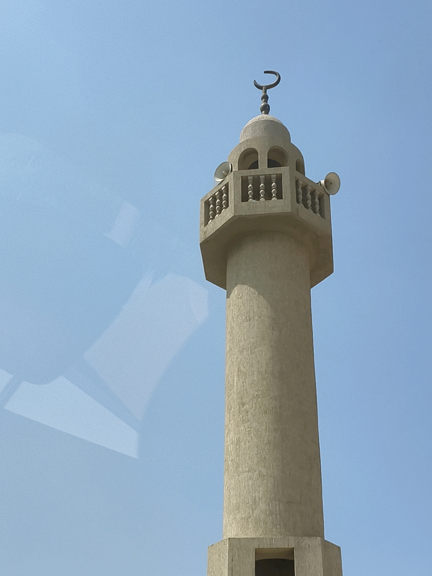 The image shows the minaret of a mosque against a clear blue sky. The minaret is made of light-colored stone and has a cylindrical body that tapers slightly towards the top. There is an octagonal section at the top of the minaret with a decorative railing. Two loudspeakers are attached to the sides of this octagonal section. At the very top of the minaret, there is a crescent moon symbol, which is a common symbol in Islamic architecture. The bottom of the minaret is partially visible, showing a rectangular opening. A faint reflection of what appears to be an indoor space is visible on the left side of the image, suggesting the photo may have been taken through a window. The minaret is tall and slender, typical of religious structures found in Dubai, United Arab Emirates.