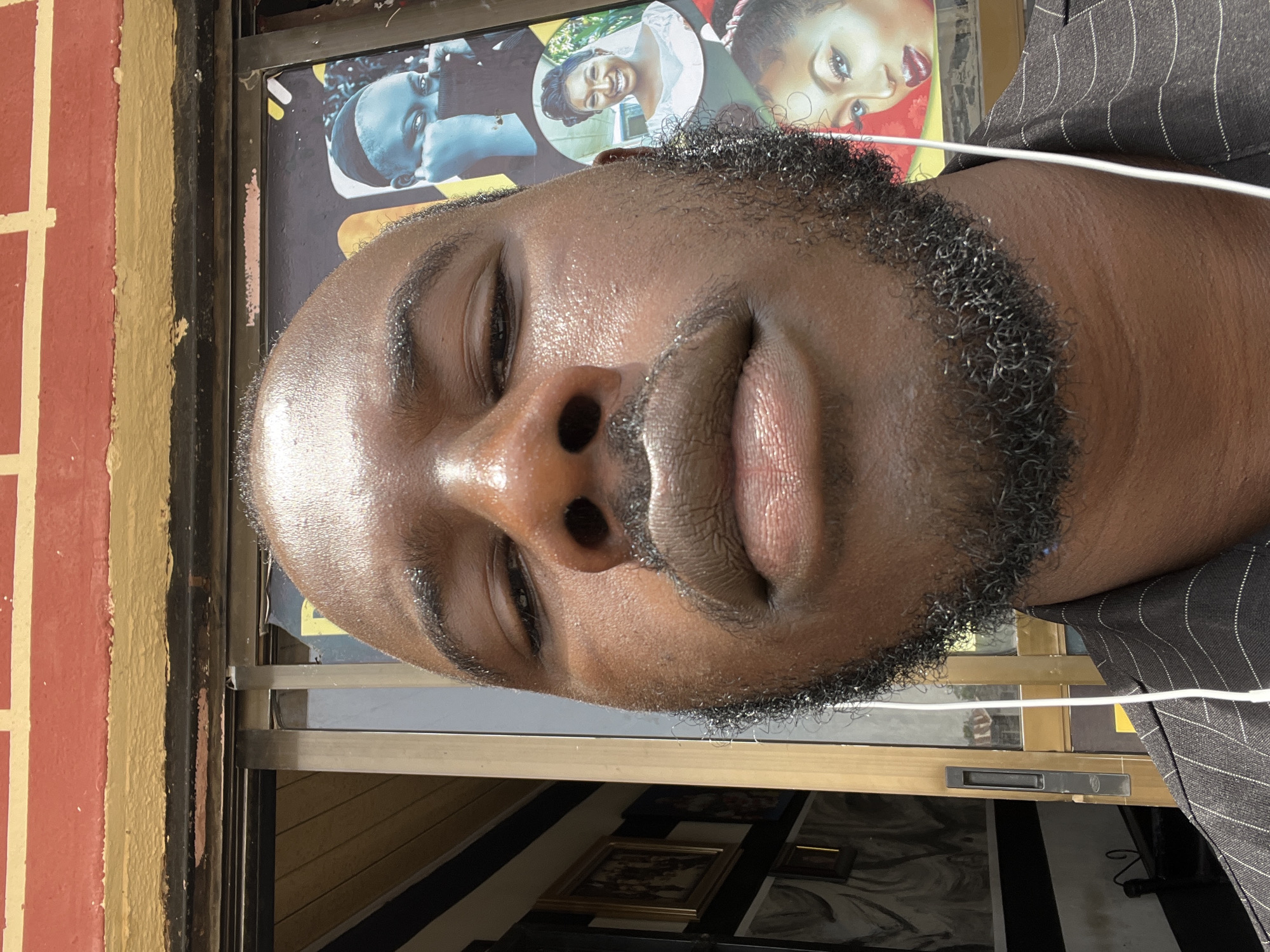 The image is a close-up, angled portrait of a Black man with a beard. His eyes are partially closed, and his skin appears to be slightly oily. He is wearing a striped suit jacket and has a white wire, likely an earbud cord, visible near his neck.
Behind him is a store window displaying posters with images of faces, possibly advertisements for a beauty salon or photography studio. The storefront is bordered by a brick and painted wall. The man is likely standing on the sidewalk outside the shop. The location is Gura Top, Nigeria. The specific environment is a commercial area. There are some gold frames visible.