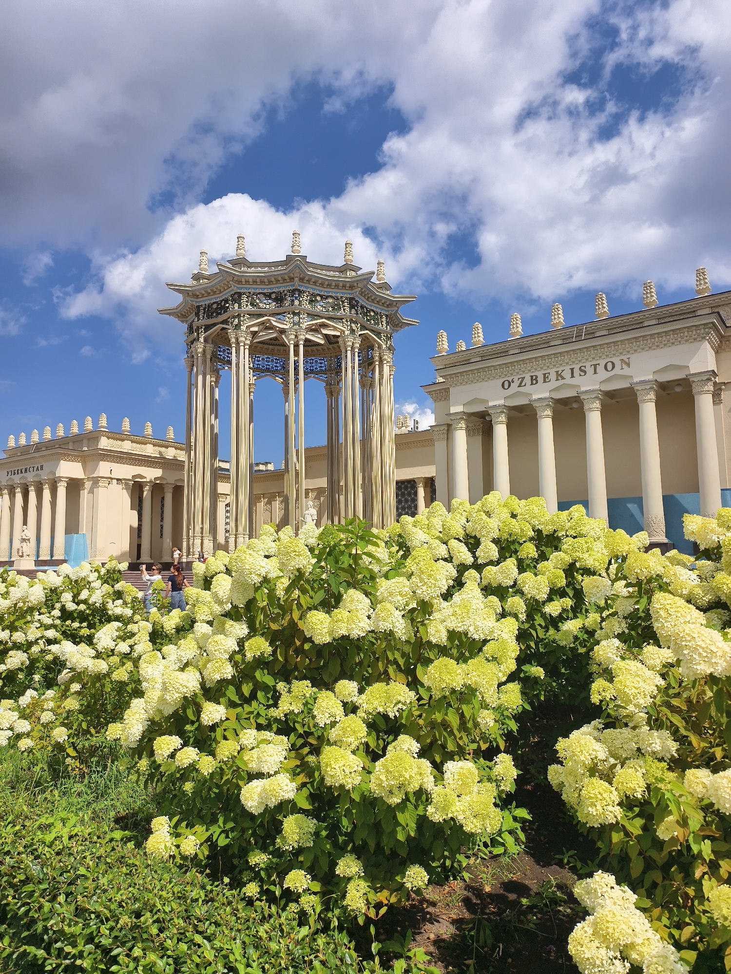 The image shows the Pavilion of Uzbekistan at VDNKh (Exhibition of Achievements of National Economy) in Moscow, Russia. In the foreground, there are lush green bushes and a large patch of blooming hydrangeas, with clusters of white and pale green flowers. Behind the flowers, a decorative, ornate pavilion with a multi-tiered roof and intricate detailing stands. To the right of the pavilion, a grand, cream-colored building with columns and the inscription "O'ZBEKISTON" is visible. The sky above is a bright blue with scattered white clouds. Two people are standing in the middle ground, near the entrance to the pavilion, with one appearing to take a photo.