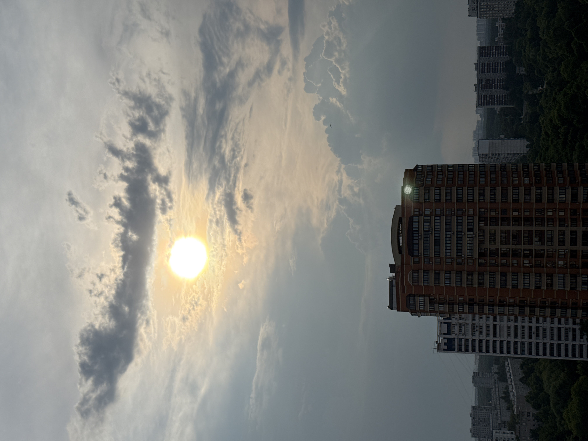 The image captures a view of the sky over a city, likely Moscow, Russia, on a day with mixed cloud cover. The sun is visible as a bright orb, partially obscured by clouds, casting a warm glow on the scene. Several high-rise buildings are present in the foreground and background, indicative of an urban environment. A large, reddish-brown apartment building with numerous windows dominates the mid-ground, with other residential buildings of varying heights and architectural styles visible further away. Lush green trees can be seen between and around the buildings, suggesting the presence of parks or residential areas with significant vegetation. The overall impression is an overcast sky with the sun attempting to break through, illuminating a cityscape.