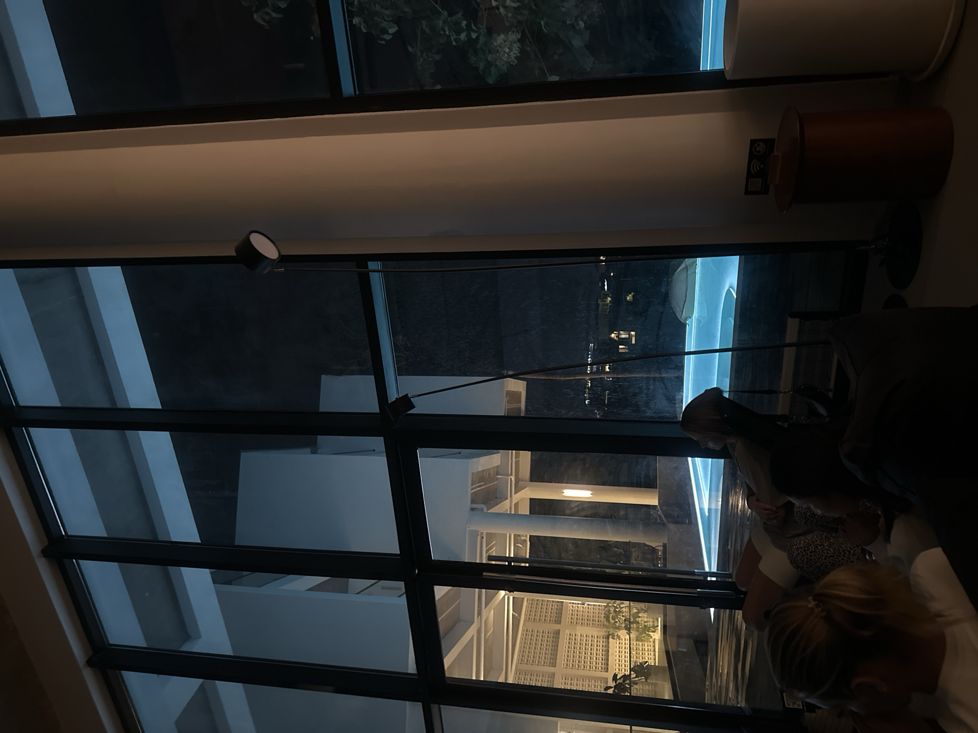 A dimly lit interior with large windows overlooking a resort's illuminated swimming pool at night. The resort appears to be in Mengwi, Indonesia. In the foreground, three individuals are seated, their faces obscured, suggesting they are observing something. One person, a woman with blonde hair tied back, faces away from the camera. Another person, also with blonde hair, is visible from the side. The third person is largely in shadow.
Beyond the seating area, a bright turquoise swimming pool is visible, its water illuminated from below. Adjacent to the pool, the resort's architecture features white pillars and a textured wall. A floor lamp with a black head is positioned near a thick white pillar in the interior space. A small, dark red cylindrical bin with a wooden top sits beside the pillar, featuring a QR code sign above it. The overall ambiance is one of relaxation and evening leisure within a modern resort setting.